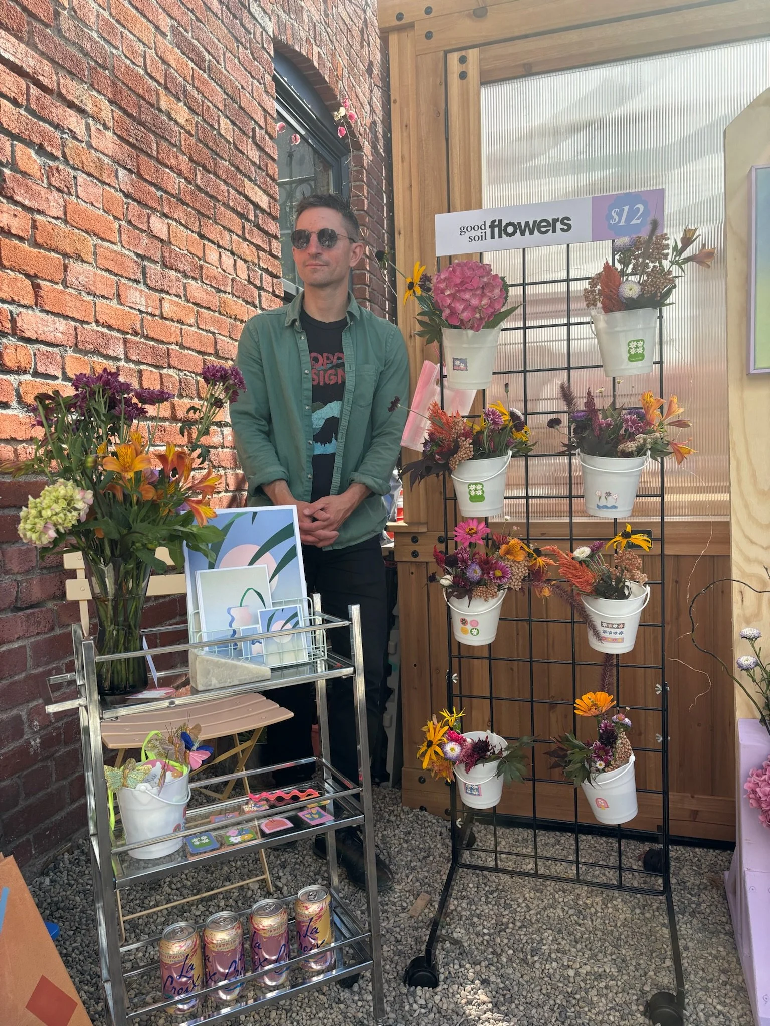 A man wearing sunglasses stands behind a display of potted flowers at a flower stall, with a sign that reads 'good soil flowers' and a price of $12. There are additional flowers in a metal cart and on a vertical rack next to him, set against a red br