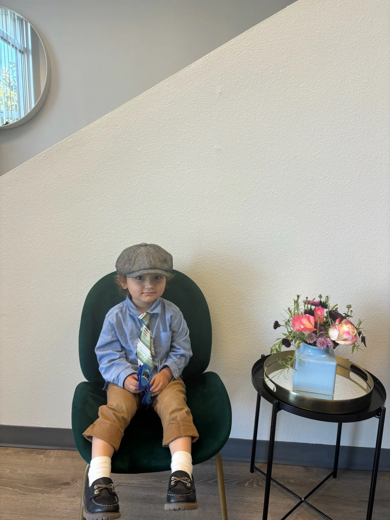 Young boy sitting in a dark green chair, wearing a grey flat cap, blue shirt, beige pants, white socks, and black shoes, holding a toy, next to a small black table with a vase of pink and purple flowers, against a light-colored wall with a round mirr