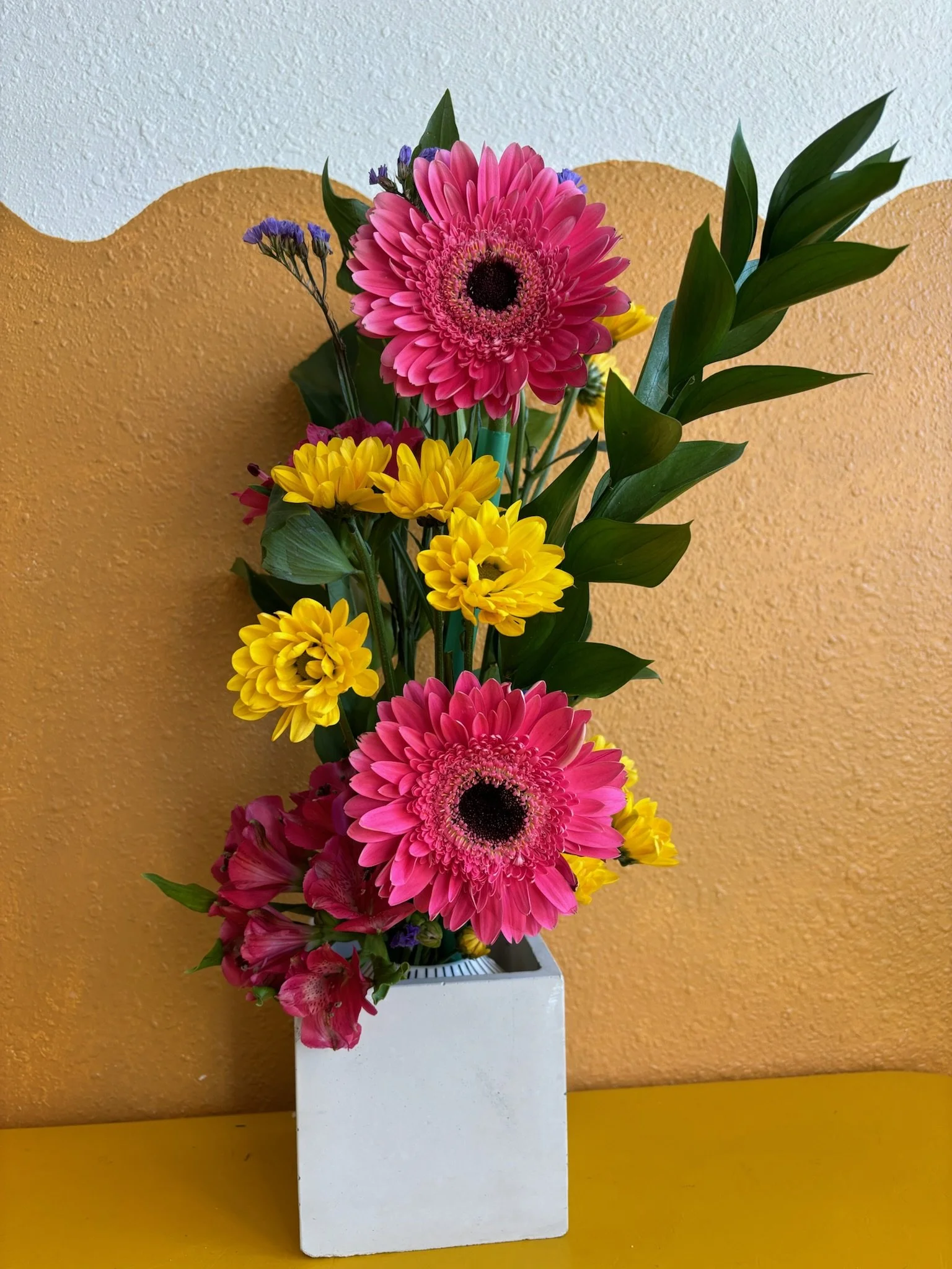 Colorful bouquet of pink, yellow, and purple flowers in a white square vase on a yellow table against a textured yellow and white wall.