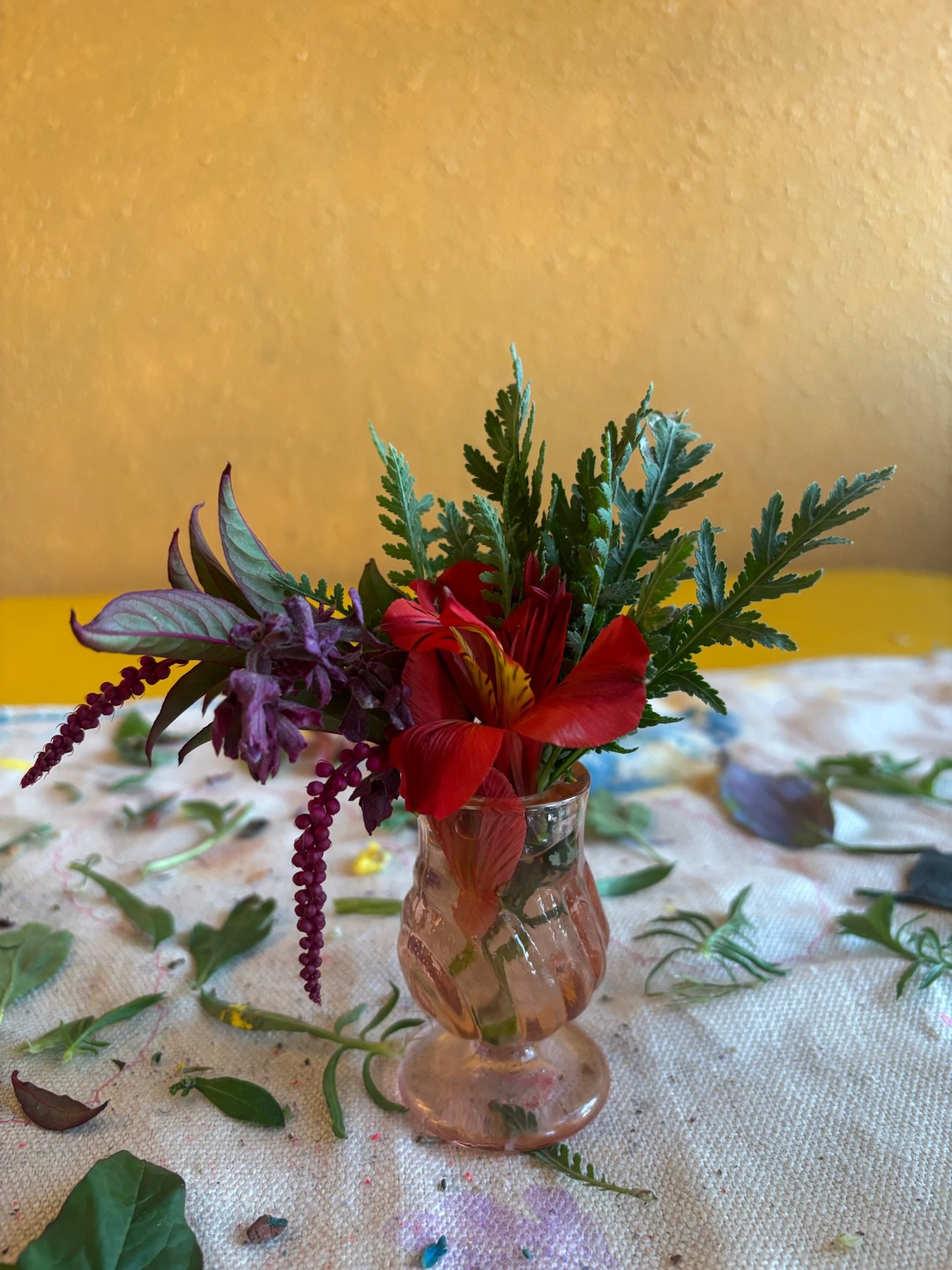 Small pink glass vase containing a floral arrangement with red, purple, and green leaves on a table with scattered leaves and flower petals, against a yellow wall background.