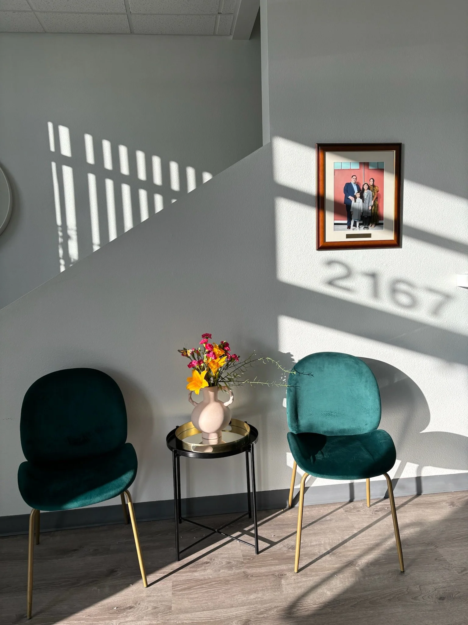 Two green velvet chairs with gold legs, a small black side table with a gold tray, a beige vase with colorful flowers, and a framed family photo on the wall with sunlight casting shadows and the room number 2167 visible.