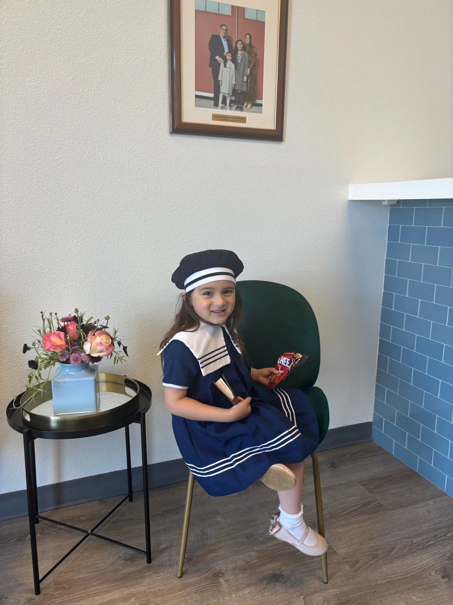 A young girl dressed in a navy blue sailor costume, sitting on a green chair inside a room, holding a snack bar and a bag of Cheetos, with a white flower arrangement on a black side table beside her and a framed family portrait hanging on the wall ab