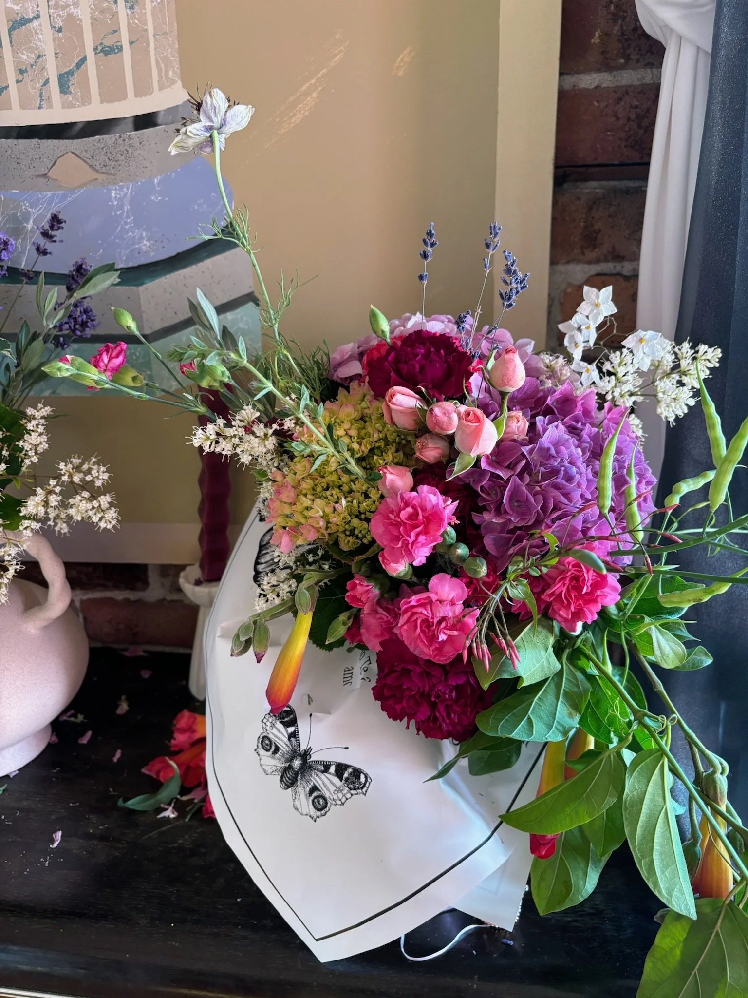 A colorful bouquet of flowers, including pink, purple, and red blooms, wrapped in white paper with a butterfly illustration, placed on a dark surface near a window.