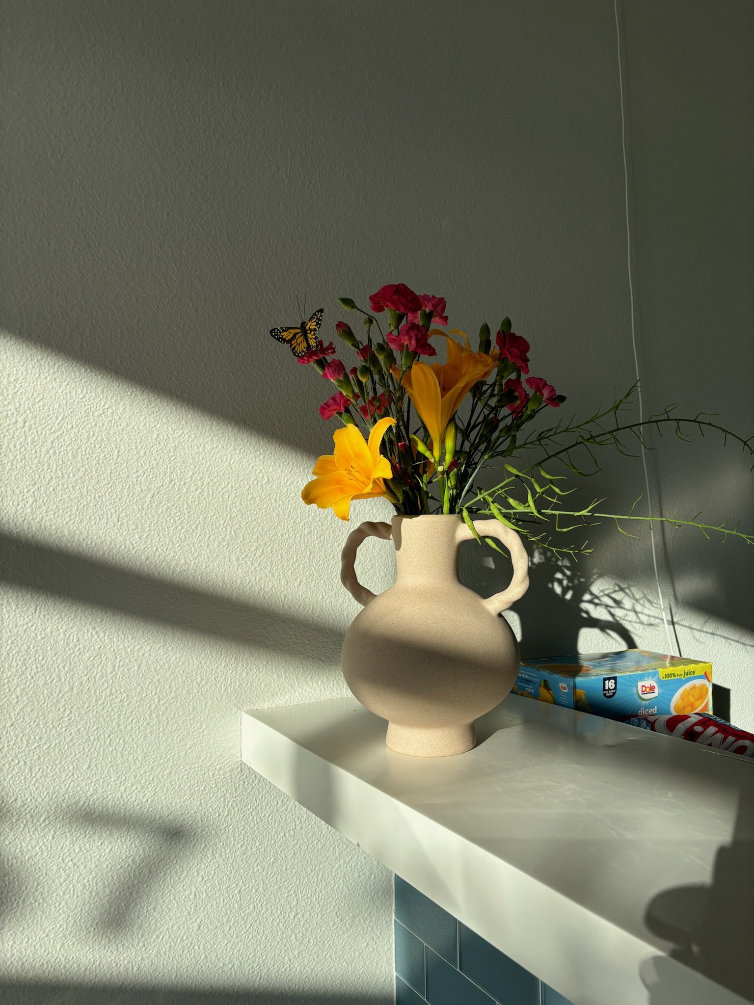 A beige ceramic vase with two handles on a white shelf, filled with colorful artificial flowers including yellow lilies and pink blossoms, with a butterfly decoration among the flowers. Sunlight casts shadows on the wall behind.
