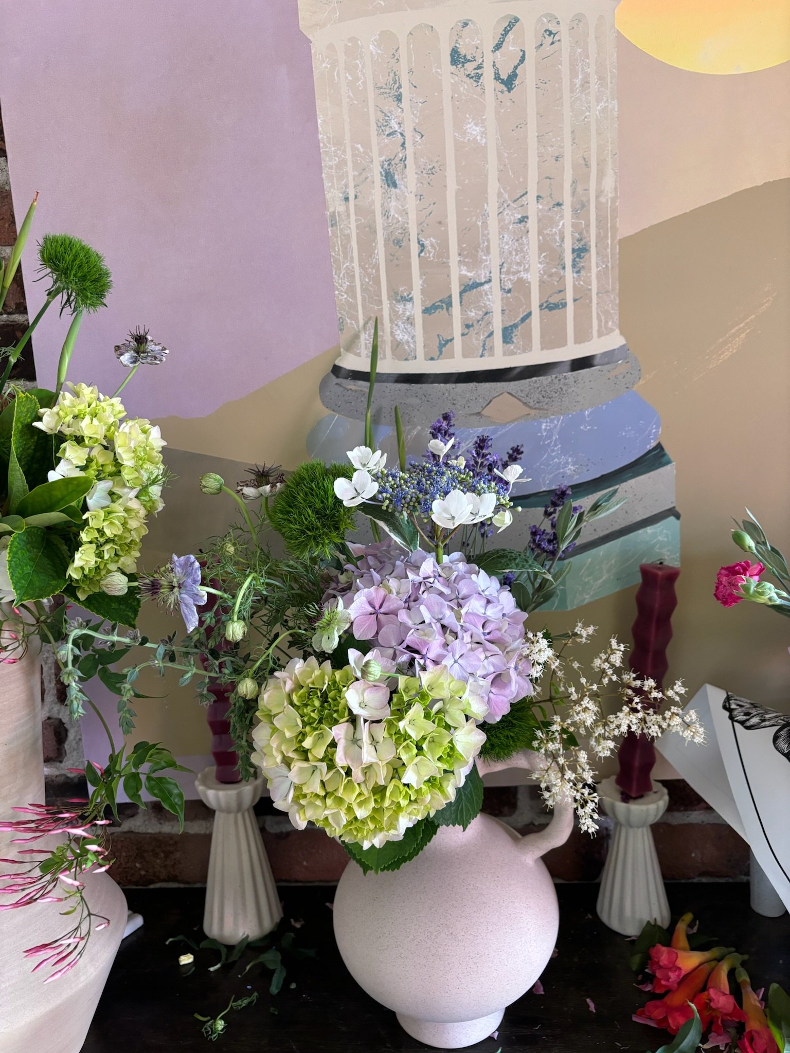 A floral arrangement with purple, white, and green hydrangeas, white daisies, lavender, and other flowers in a round, light-colored vase on a dark surface, with abstract artwork and candles in the background.