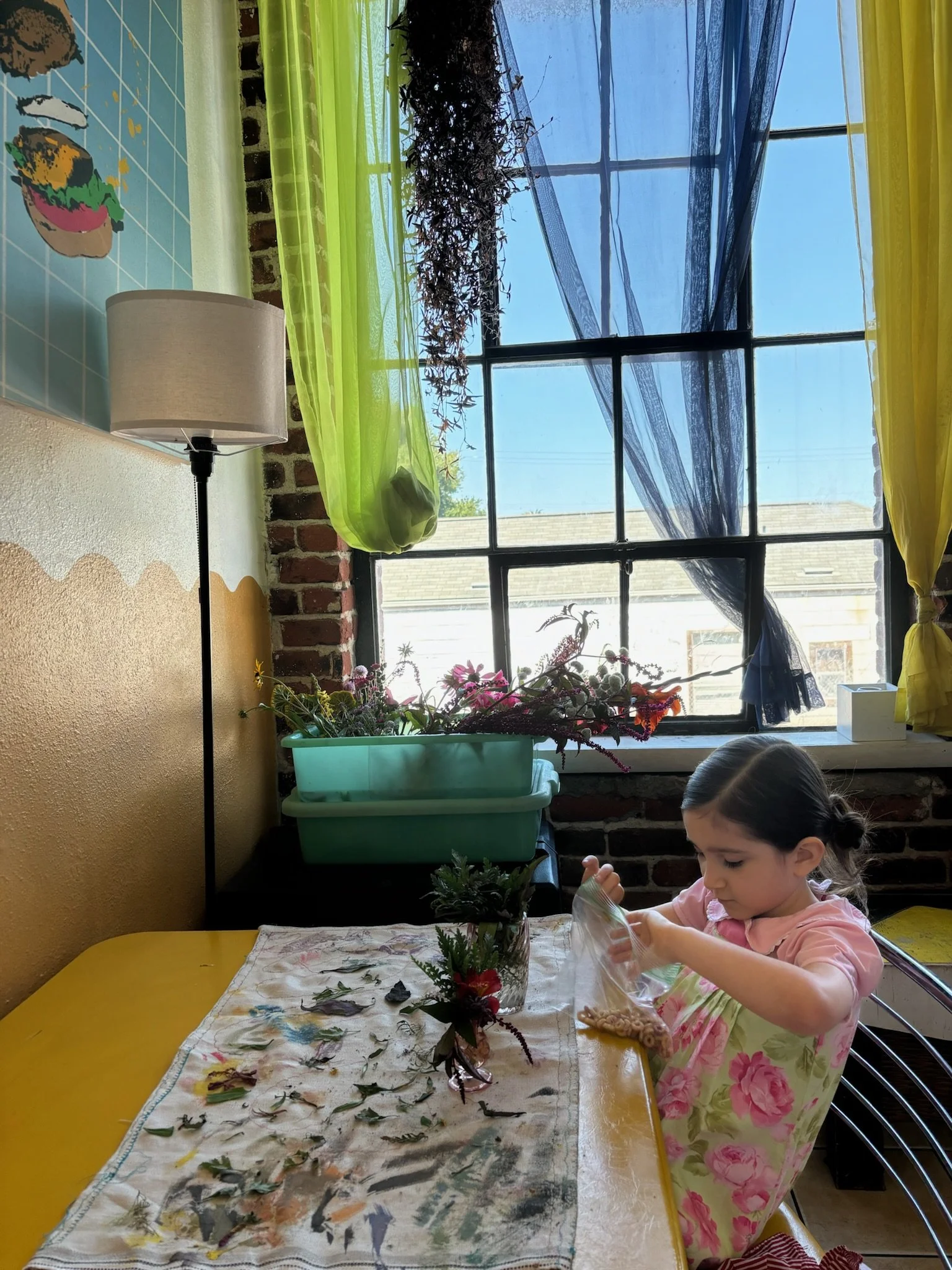 A young girl with dark hair in a ponytail sits at a yellow table, decorations and leaves are spread on the table, and she is putting cereal into a plastic bag. There is a window with blue and yellow curtains, and the window has plants hanging by it, 