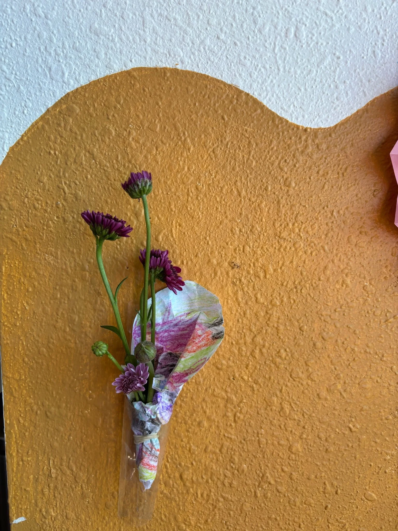 A bouquet of purple flowers wrapped in colorful paper is mounted on an orange textured wall.