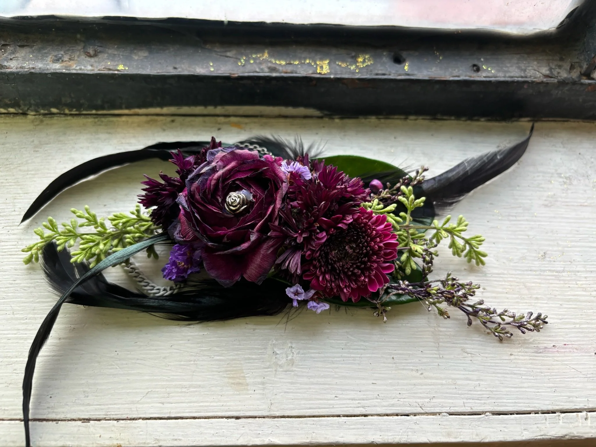 A floral corsage with dark purple flowers, green leaves, and black feathers, resting on a white windowsill.