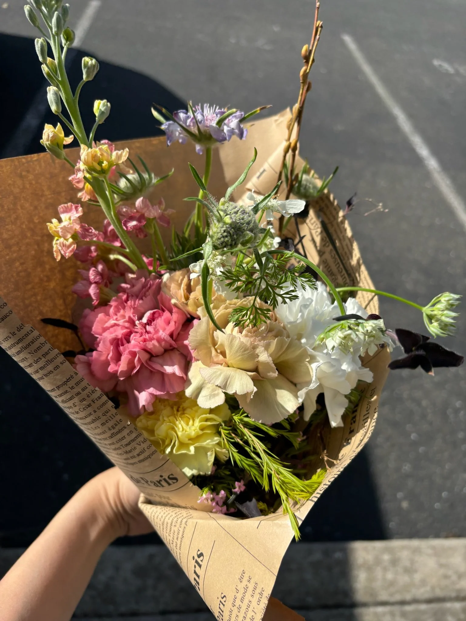 Hand holding a bouquet of mixed flowers wrapped in newspaper paper, with a dark pavement background.