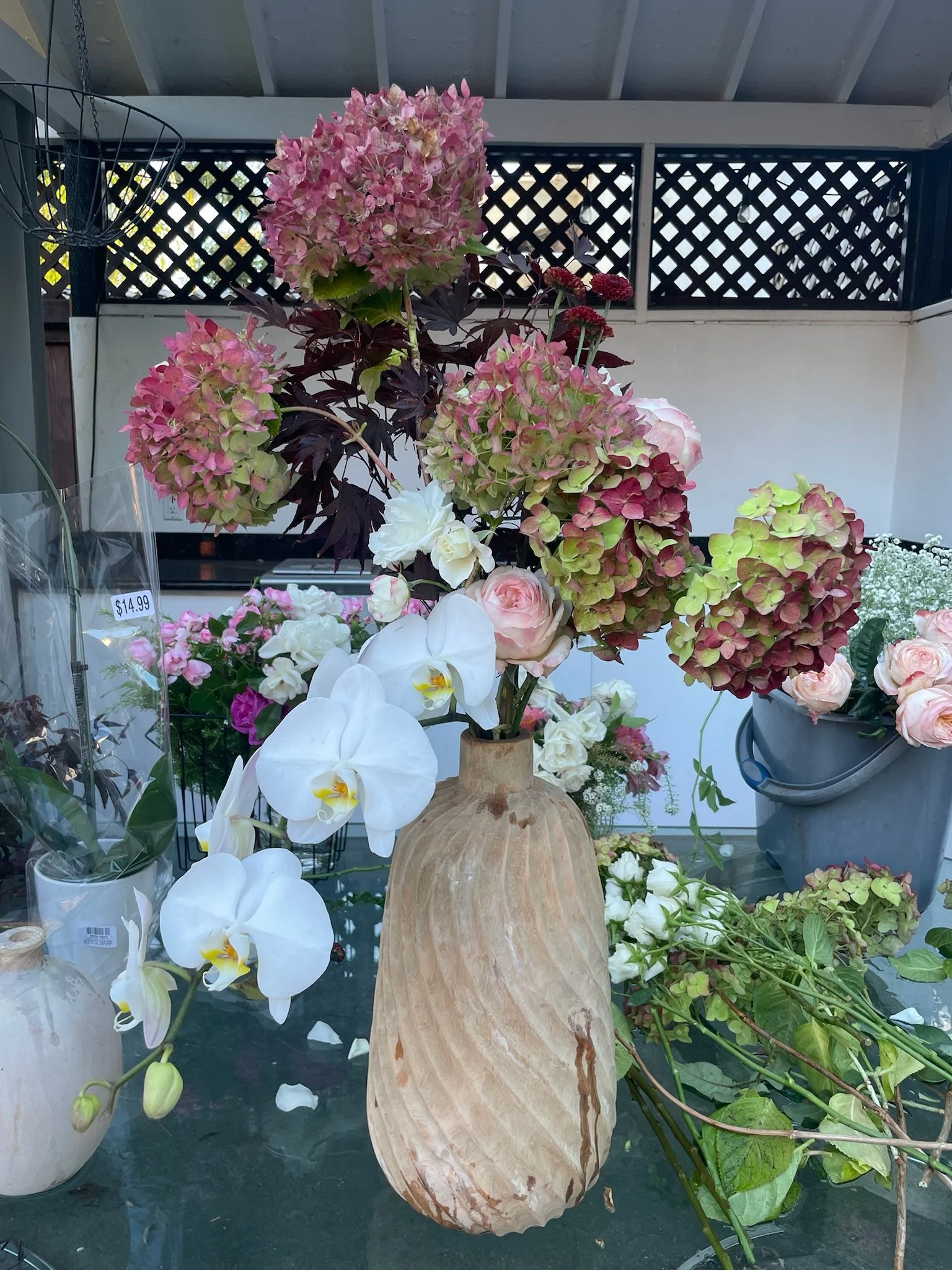 A large bouquet of pink and green hydrangeas, white orchids, and pale pink roses arranged in a rustic wooden vase on a glass table at a flower shop in Alameda, CA.