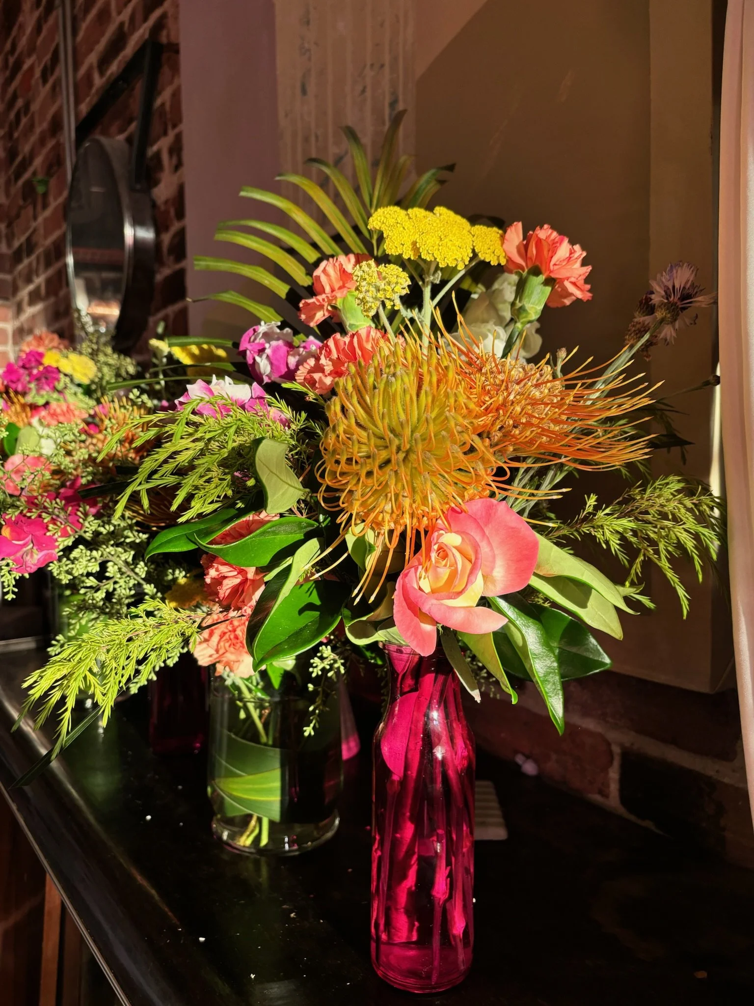 A colorful bouquet of flowers in a tall pink glass vase on a black surface, with a mirror and brick wall in the background.