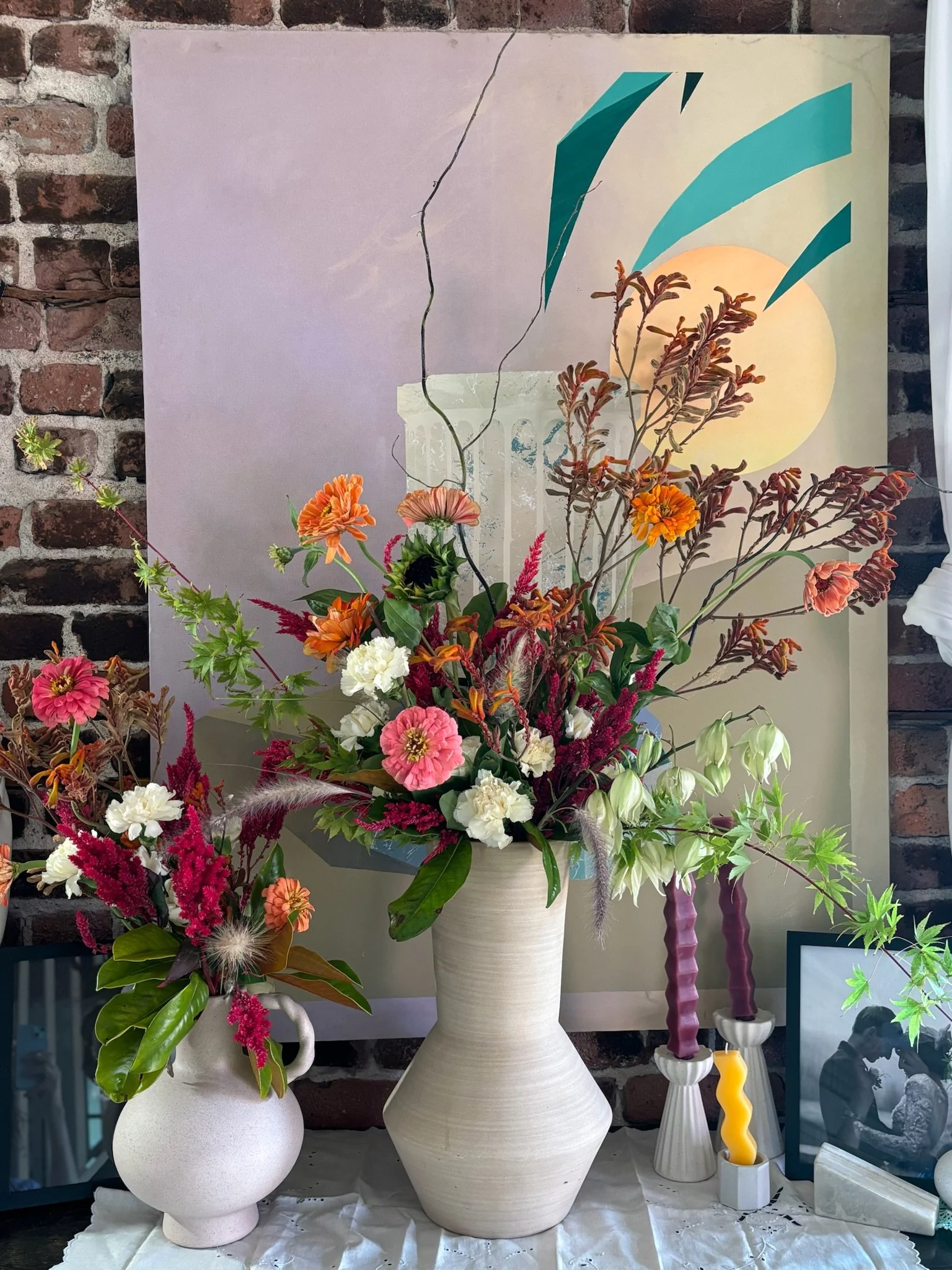 A large bouquet of colorful flowers in a white vase on a table, with a smaller flower arrangement beside it, against a brick wall with an abstract painting and framed photo in the background.