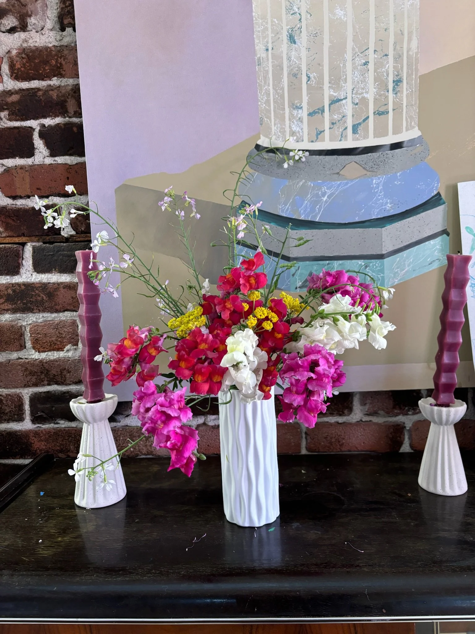 A floral arrangement with pink, white, and yellow flowers in a white vase, flanked by two white candlesticks with pink candles, on a dark wooden surface against a brick wall and abstract painting.