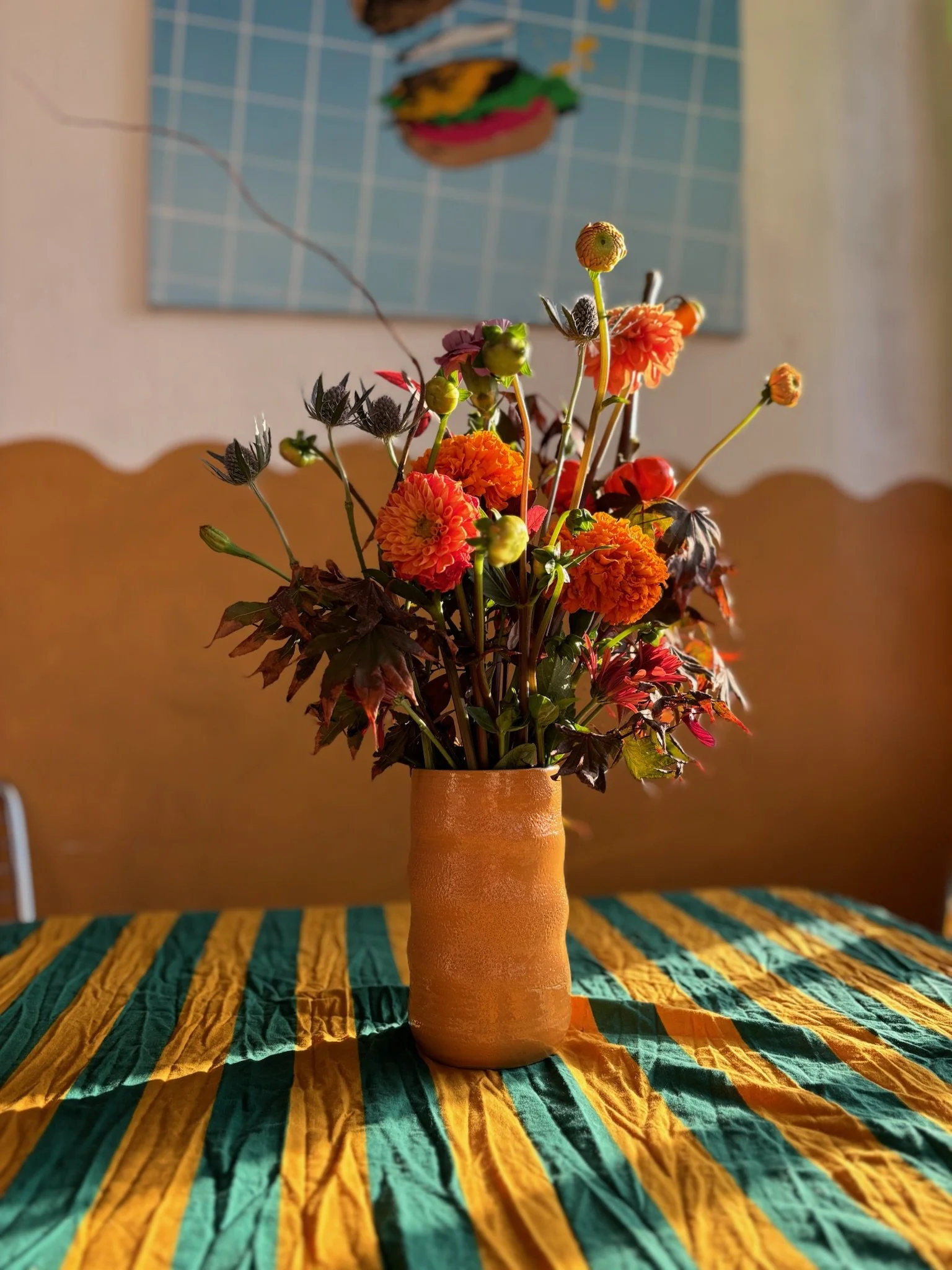A bouquet of orange and pink flowers in a terracotta vase on a yellow and green striped tablecloth, with a blue grid wall and framed picture in the background.