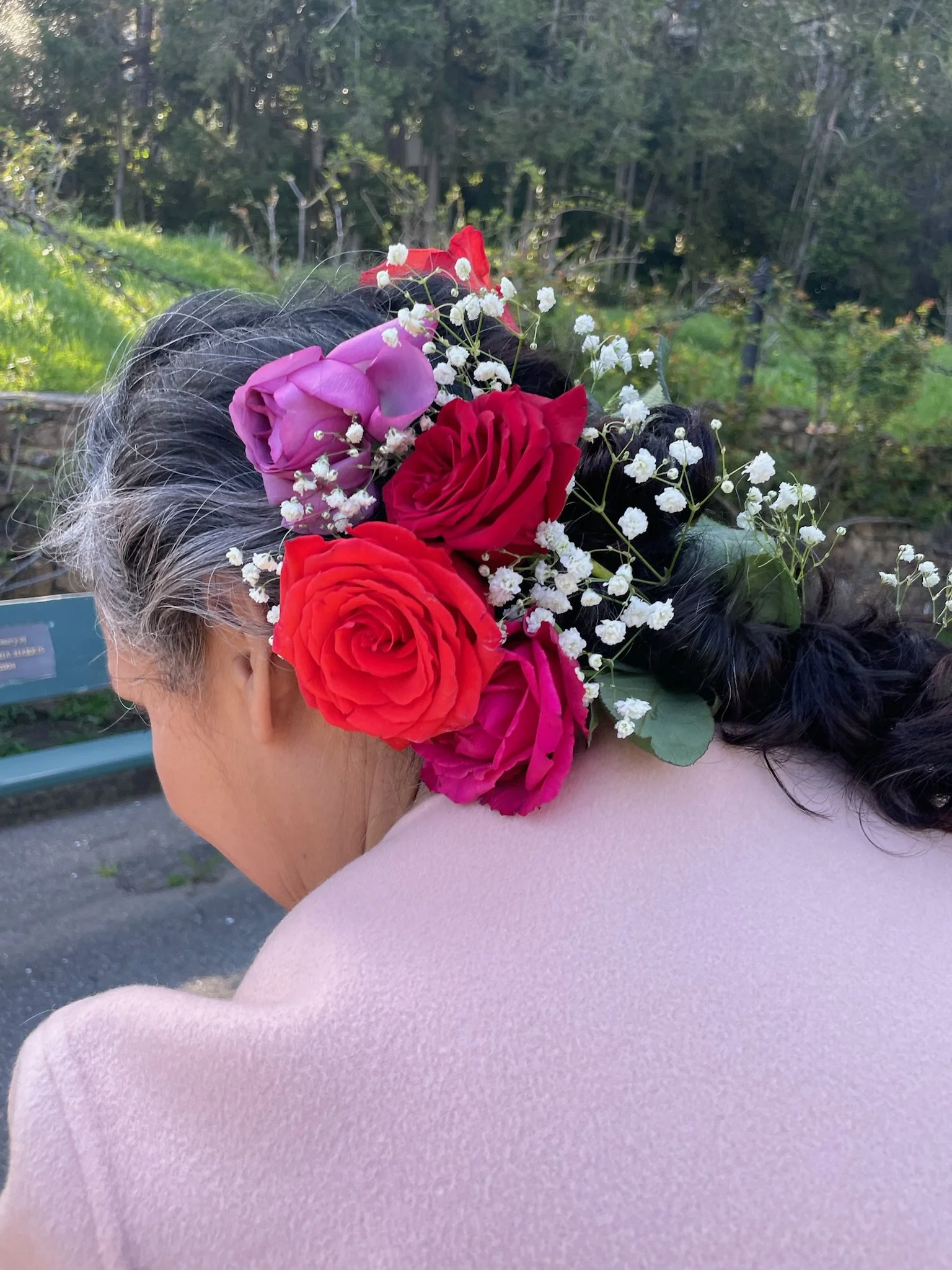 Side view of an elderly woman with gray hair wearing a pink blazer, decorated with a floral headpiece consisting of pink, red, and purple roses, and small white flowers, in an outdoor garden setting in Oakland, CA.