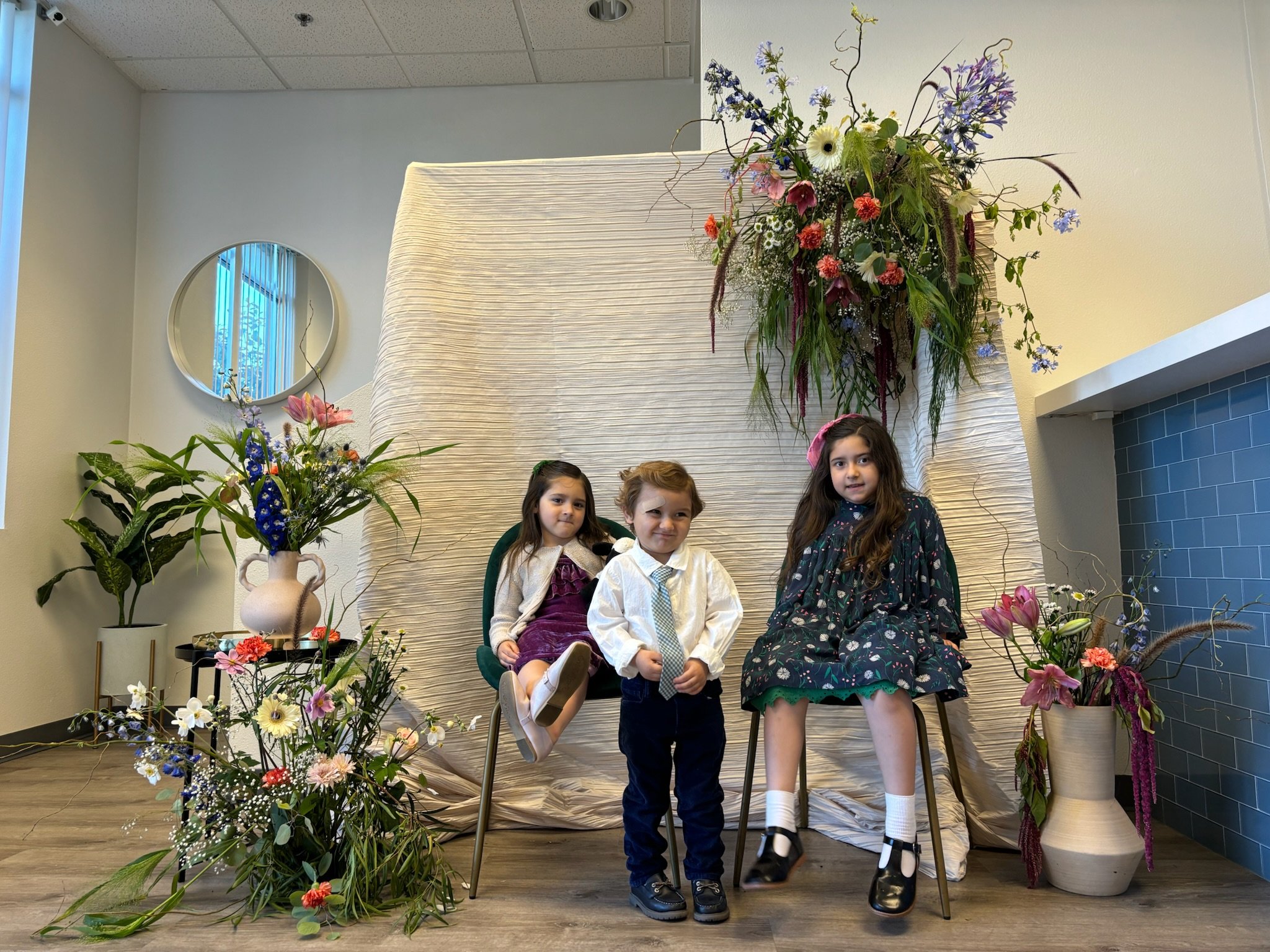 Three children in formal attire pose in front of a floral backdrop with large flower arrangements and a textured white fabric backdrop.