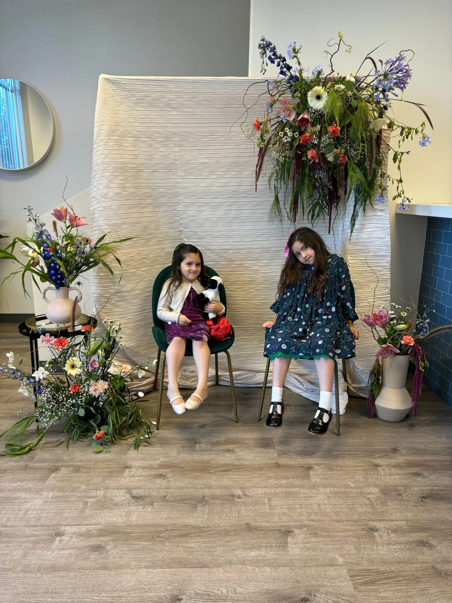 Two young girls sitting on chairs in front of a floral backdrop, surrounded by vases of flowers and greenery.