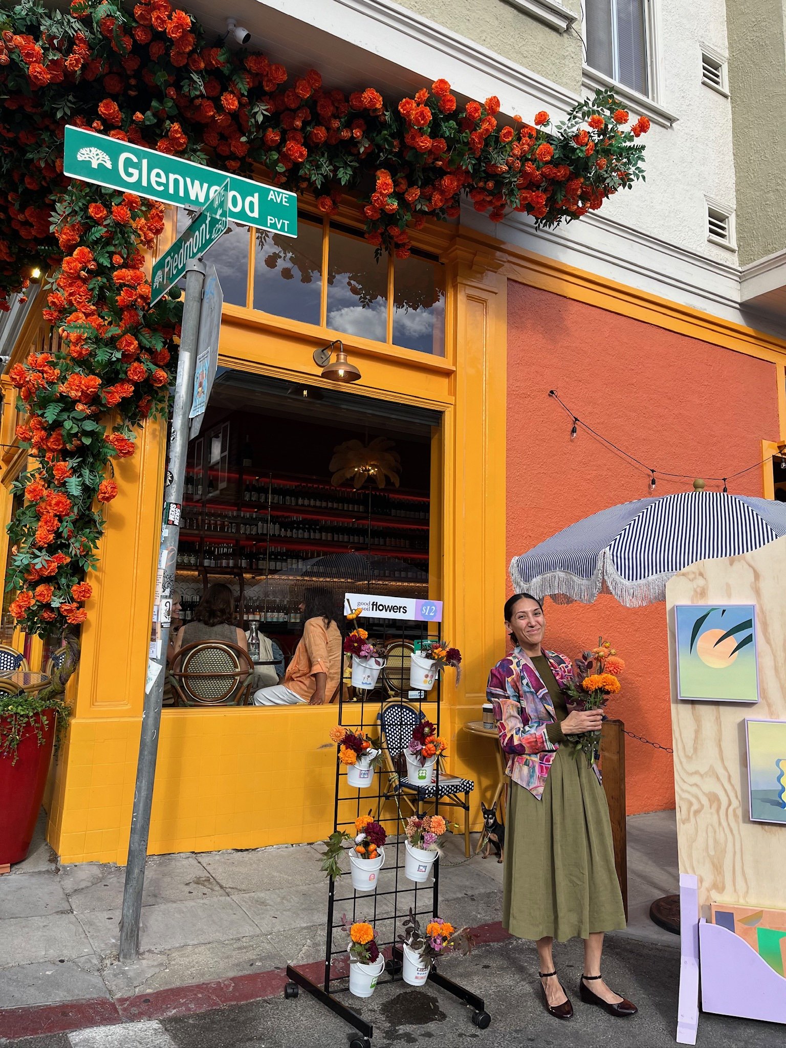 A woman holding a bouquet of orange and purple flowers standing outside a colorful flower shop on a street corner, with a sign indicating the intersection of Glenwood Avenue and Piedmont Avenue.