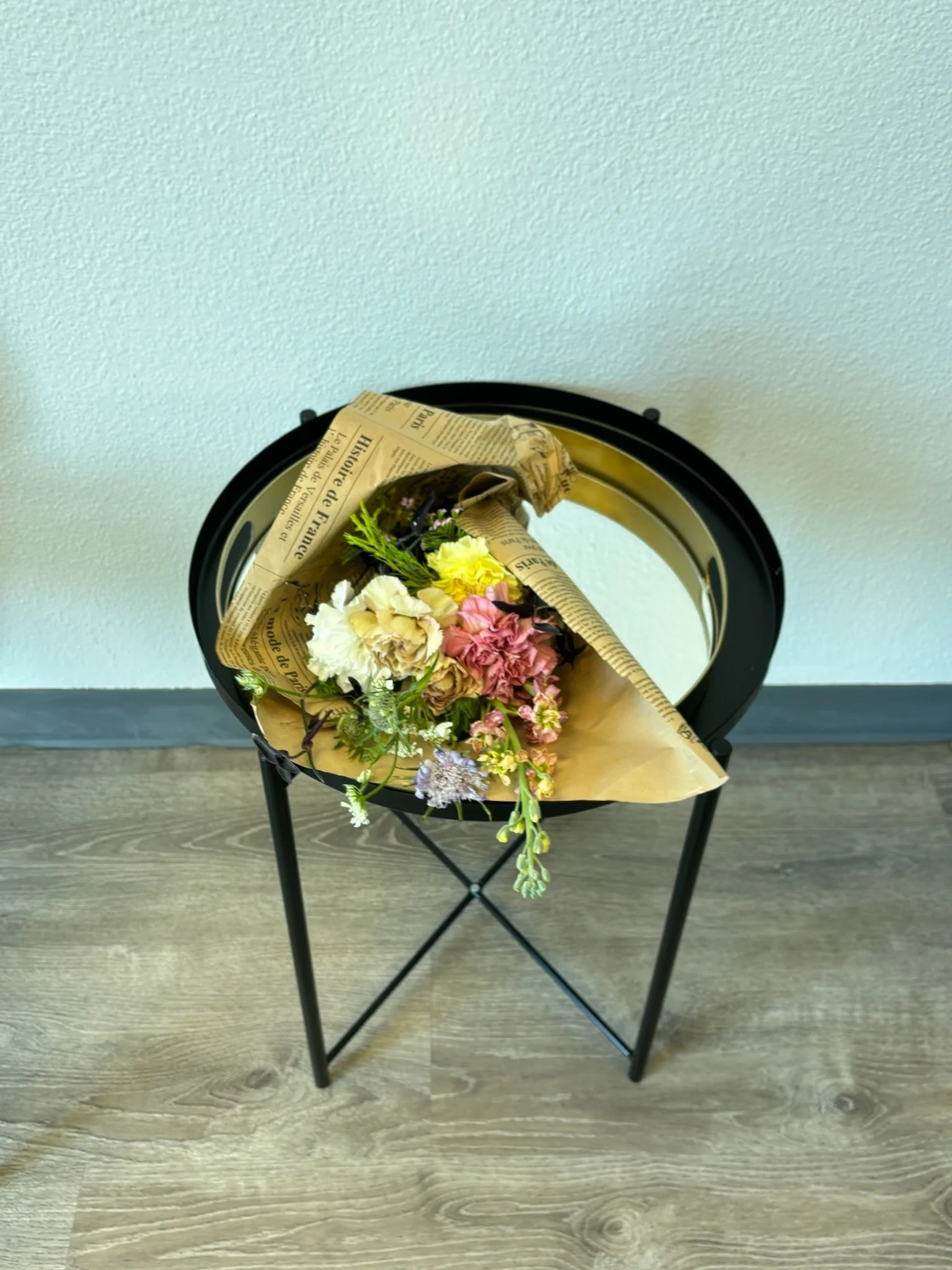 A bouquet of mixed flowers, wrapped in newspaper and kraft paper, resting on a small black metal stand against a plain white wall.
