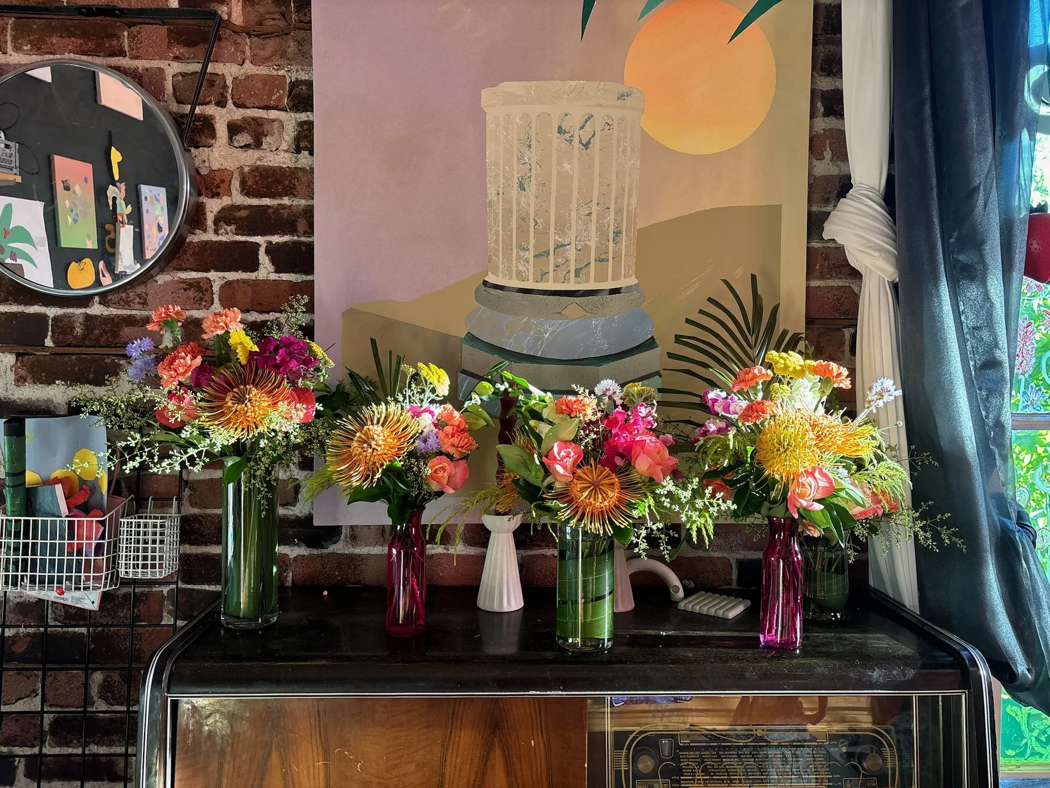 Three colorful flower arrangements in vases on a black surface, with a brick wall, artwork, and curtains in background.