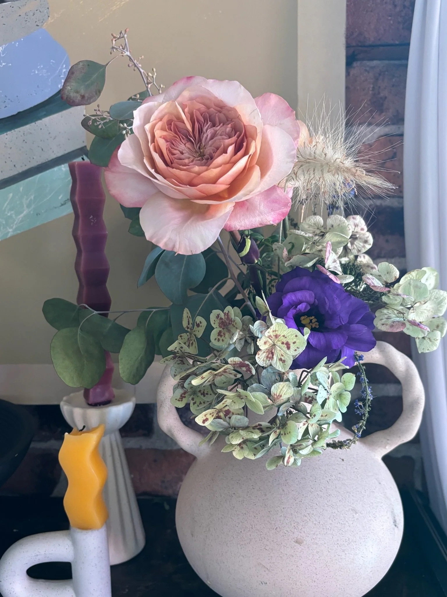 A beige ceramic vase with pink, purple, and green flowers and greenery, placed near candles and decorative items.