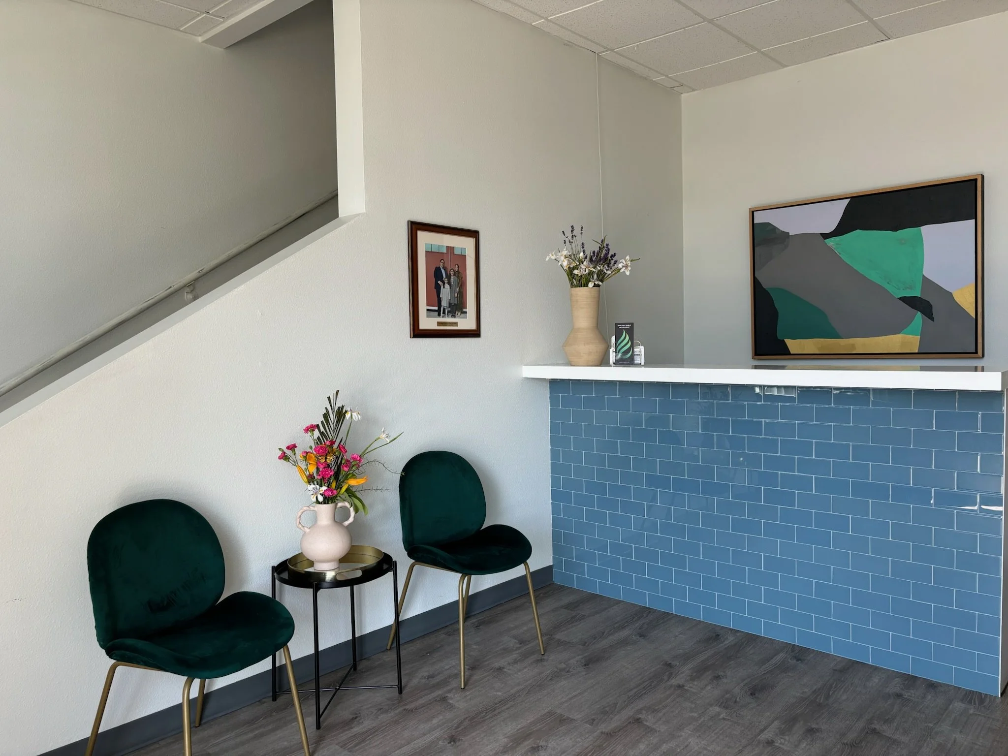 Interior of a waiting area with two green velvet chairs, a small black side table with a pink flower arrangement, framed photos and paintings on the white wall, and a blue tiled reception counter with a white top and decorative items on it.