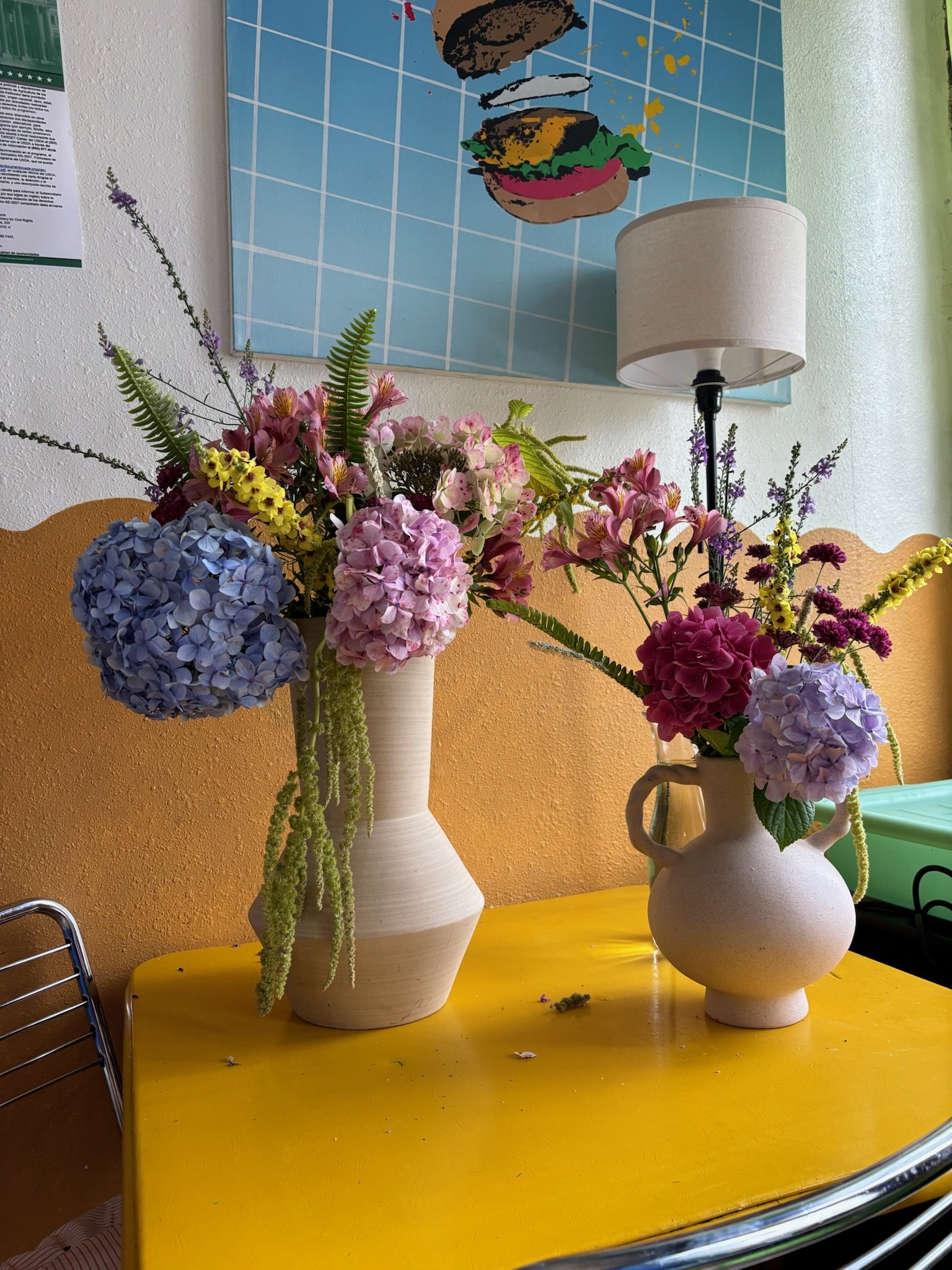 Two vases with colorful flower arrangements on a yellow table, with a framed artwork of a burger behind them and a white lamp beside the artwork.
