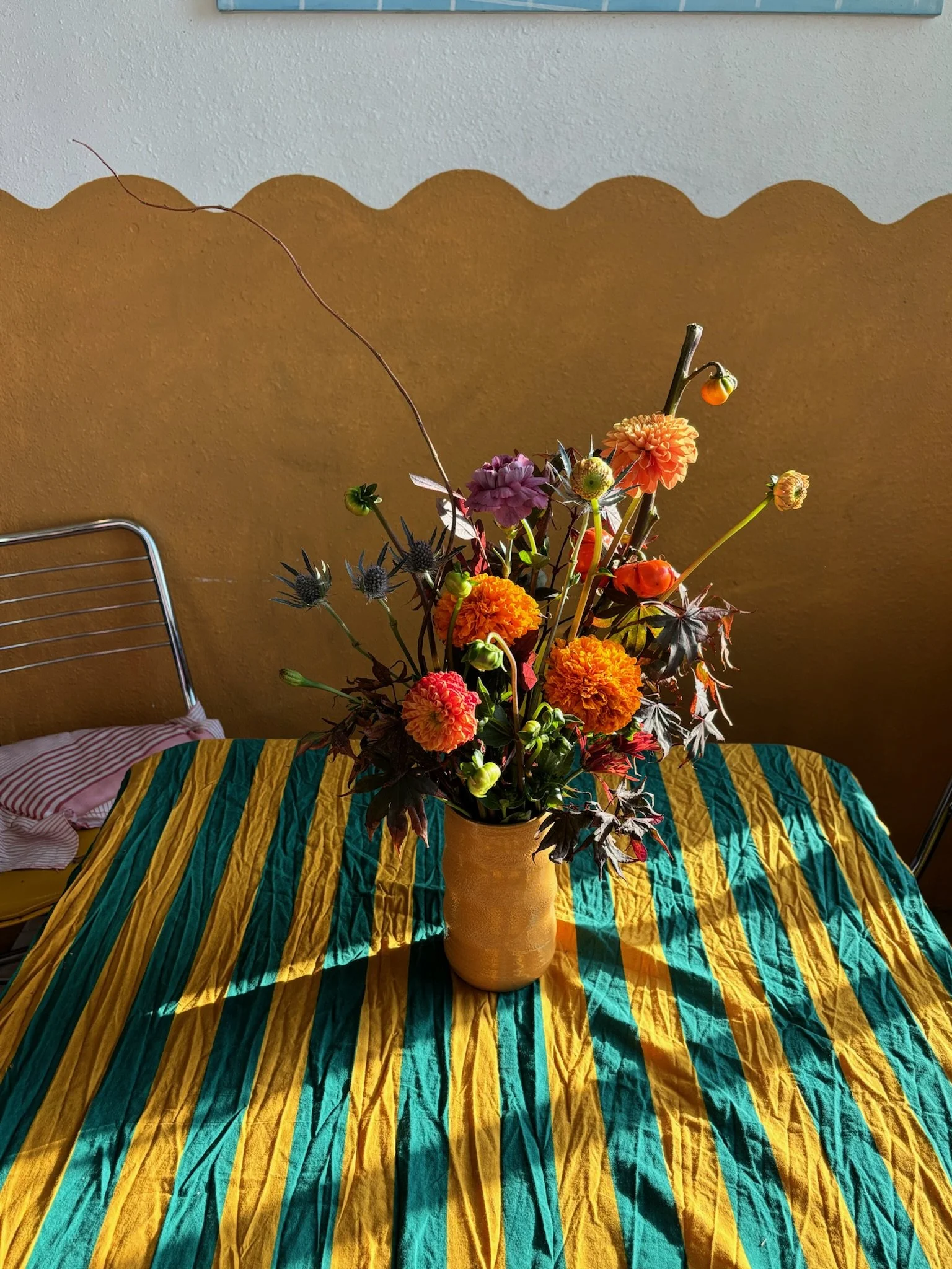 A vase of colorful flowers, including marigolds and dahlias, placed on a striped tablecloth with yellow and teal stripes, on a table with sunlight casting shadows.
