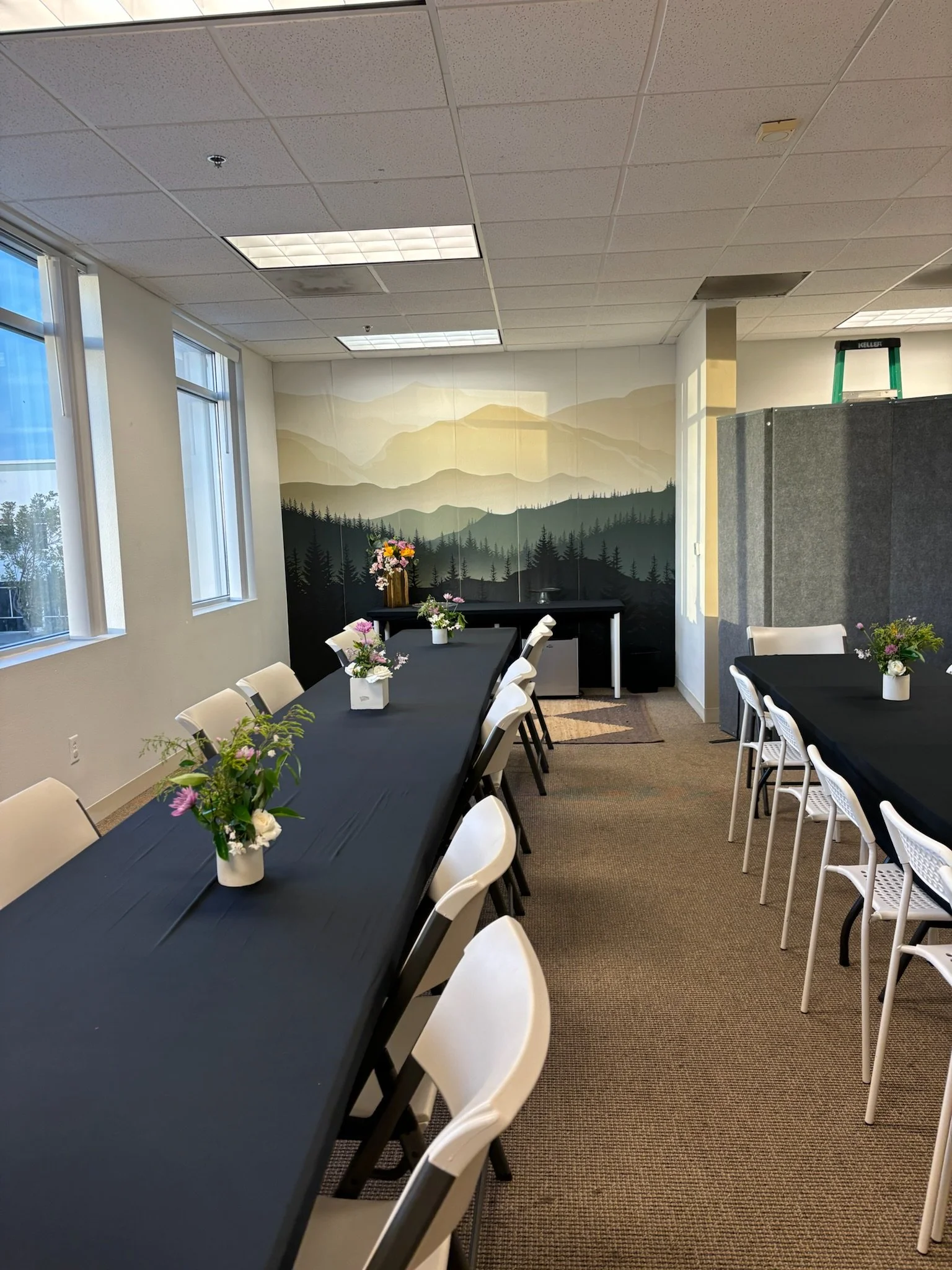 Indoor dining area with long black tables, white chairs, and floral centerpieces. There is a mountain landscape mural on the back wall, and windows on the left side letting in natural light.