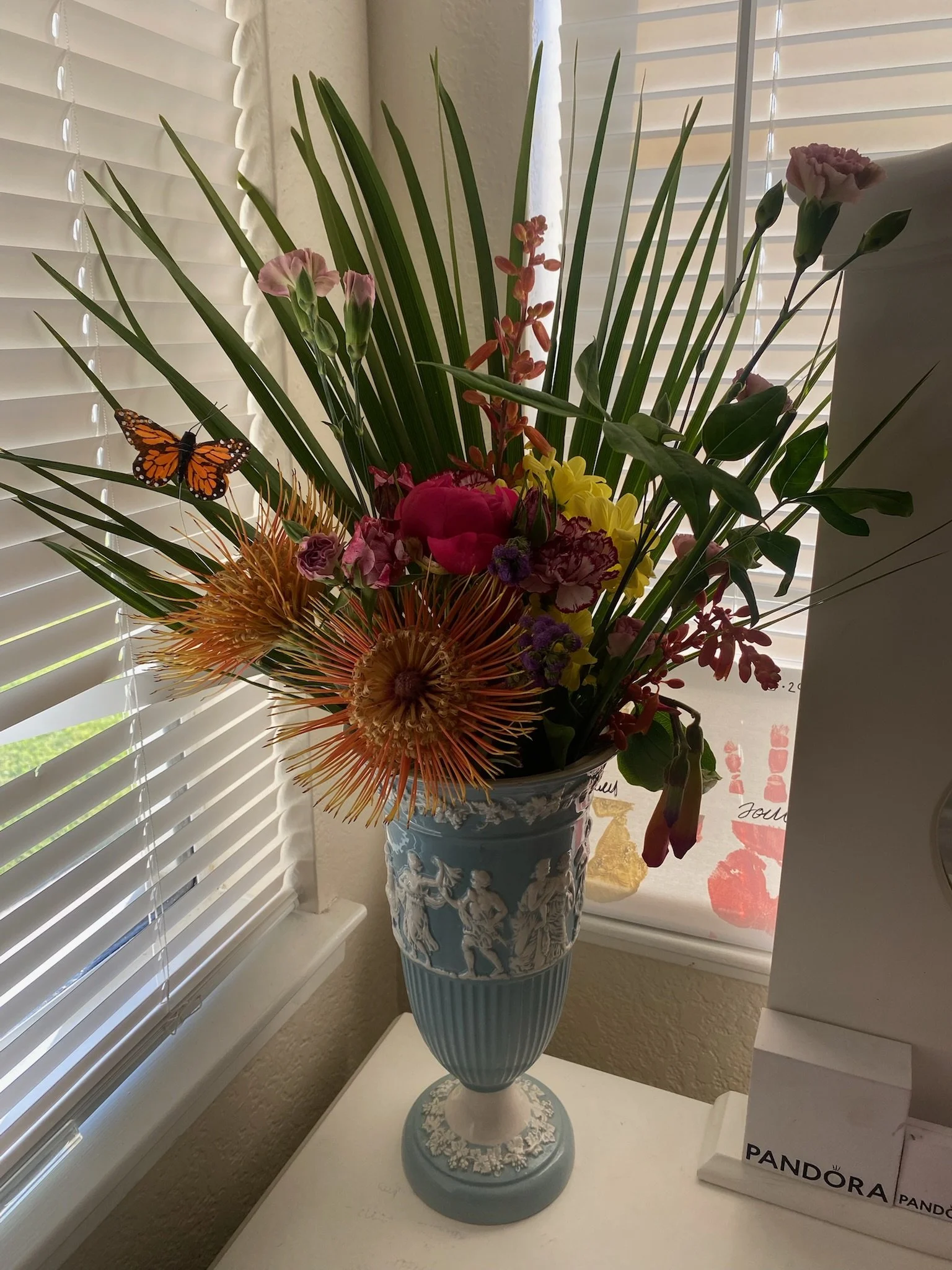 A decorative bouquet of various colorful flowers in a blue vase with classical figures design, placed on a white table near window blinds.