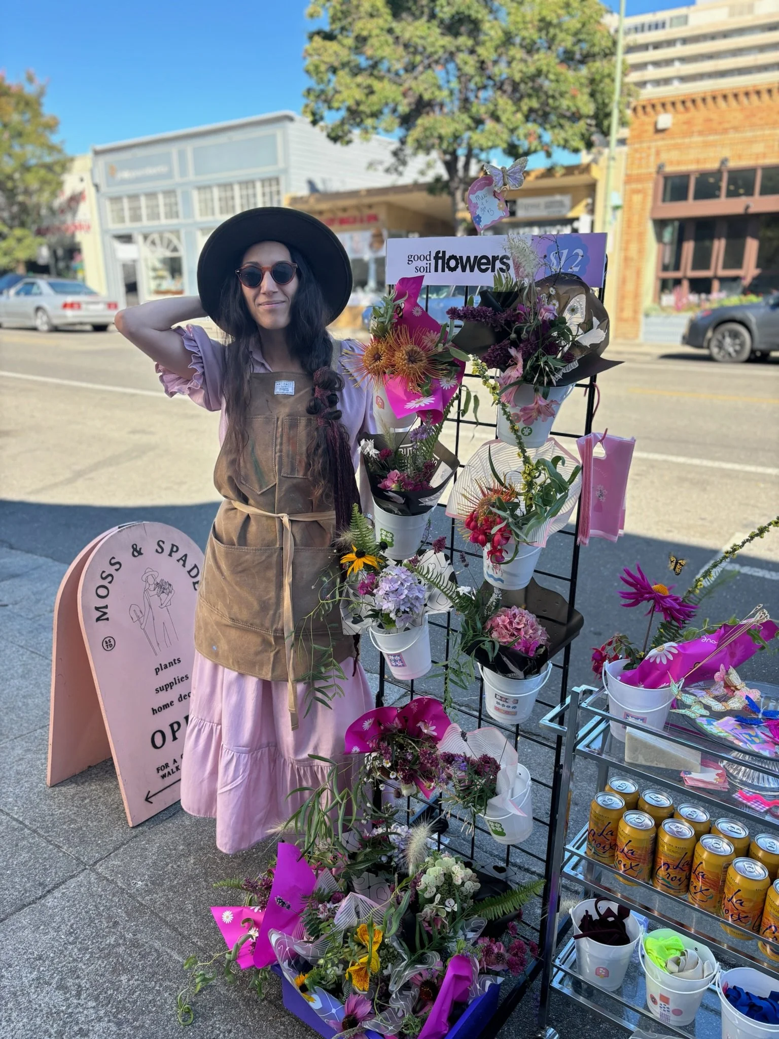 A woman with long dark hair, wearing a black wide-brimmed hat, sunglasses, a pink long skirt, and a brown apron, stands on a sidewalk next to a display of colorful flower bouquets in white buckets and pink wrapping. She is positioned beside a sign fo