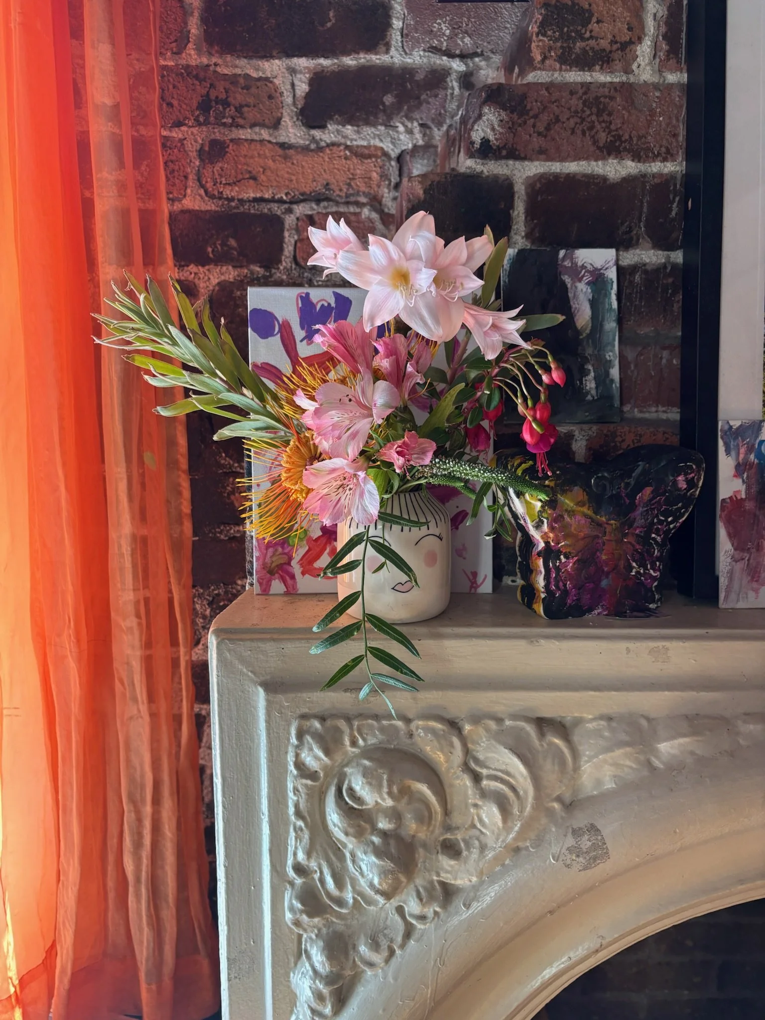 A floral arrangement with pink lilies, pink and yellow flowers, and greenery in a white face-shaped vase, placed on an ornate white fireplace mantle against a brick wall, with colorful artwork and decorative objects nearby.