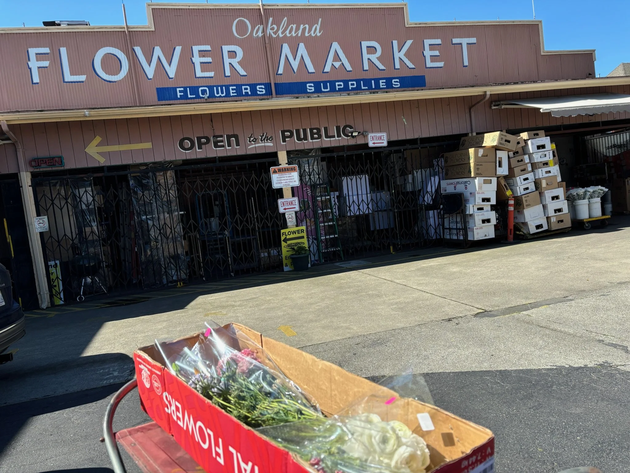 The exterior of Oakland Flower Market with stacked flower boxes and supplies outside. A cardboard box with fresh flowers is in the foreground, and signs indicating the store is open to the public and accepts flowers are visible.