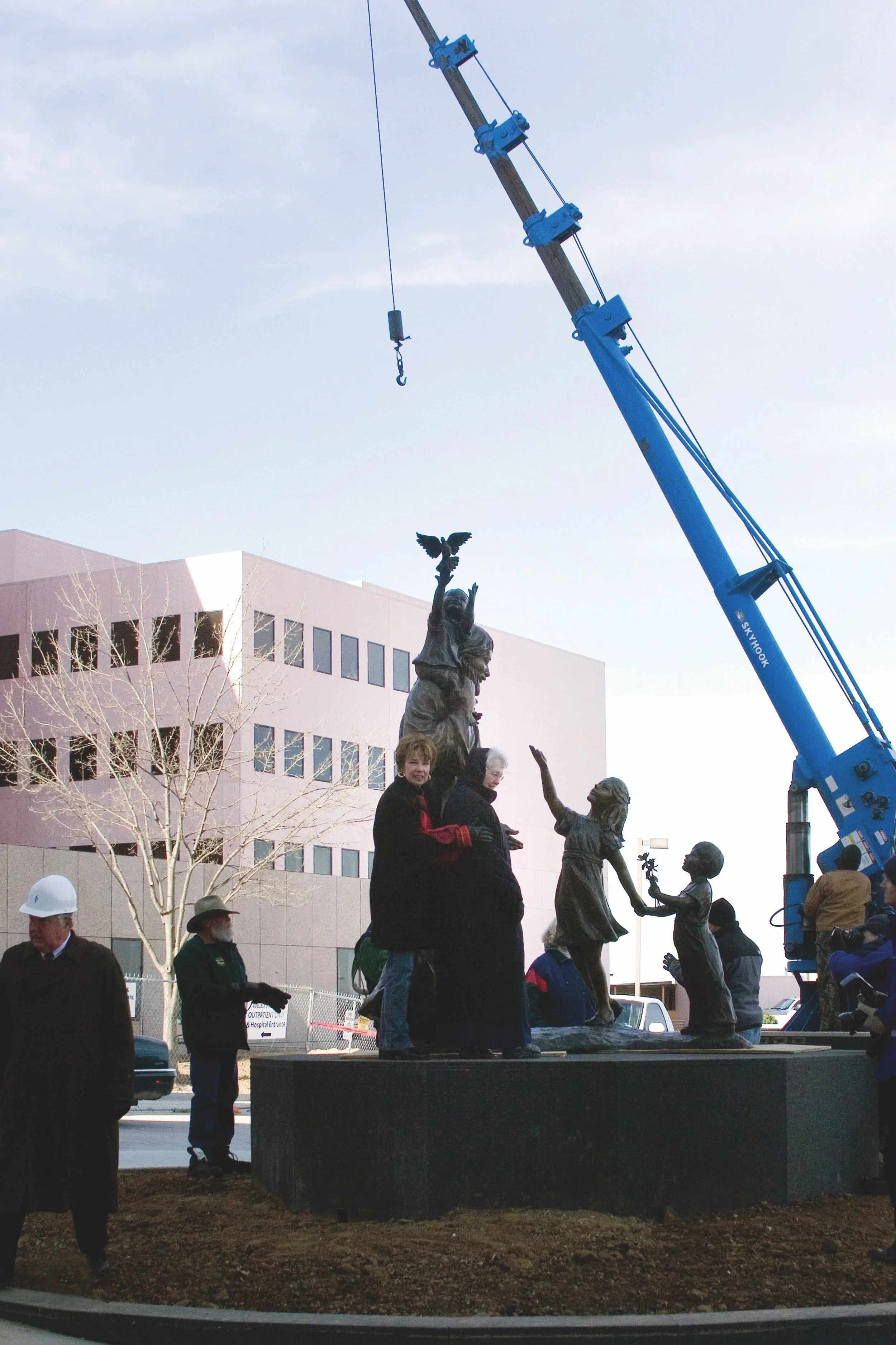 A bronze statue of children and a woman with a bird on a pole, being installed with a crane outside a modern building.