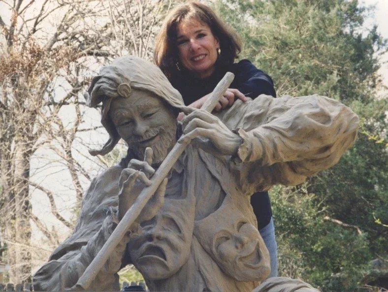 Rosalind Cook posing with a detailed clay sculpture of a fantasy character with multiple faces, holding a staff, outdoors with trees in the background.