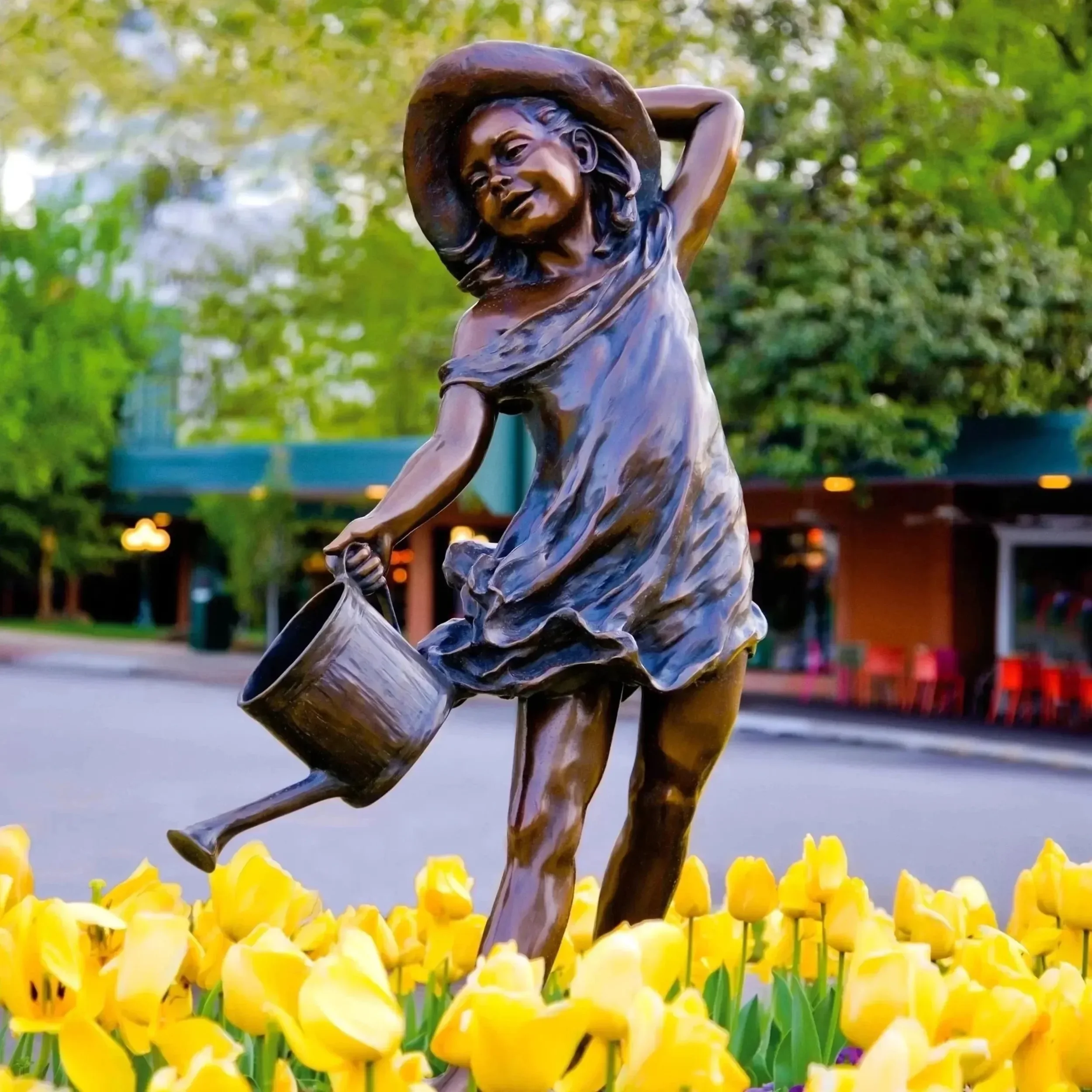 Bronze statue of a girl watering yellow tulips in a park with trees and buildings in the background.