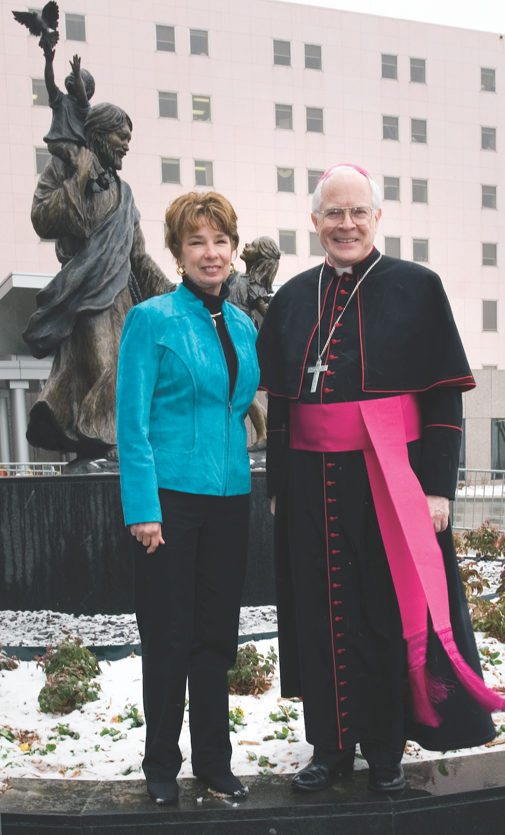Rosalind Cook wearing a blue jacket and a bishop in religious robes stand together in front of a sculpture with a modern building in the background.