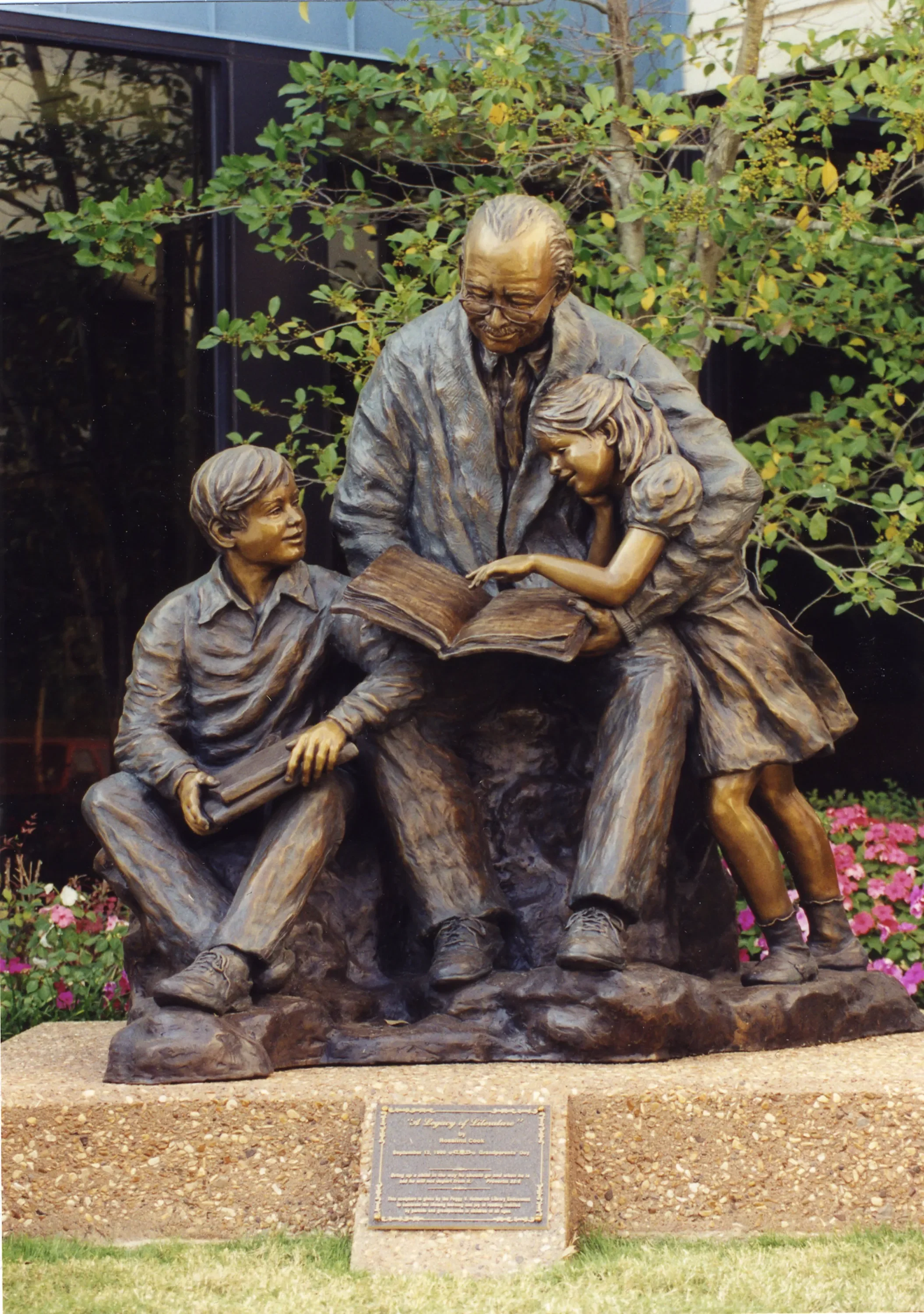 Bronze statue of an elderly man showing two children a book outdoors, surrounded by flowers and bushes.