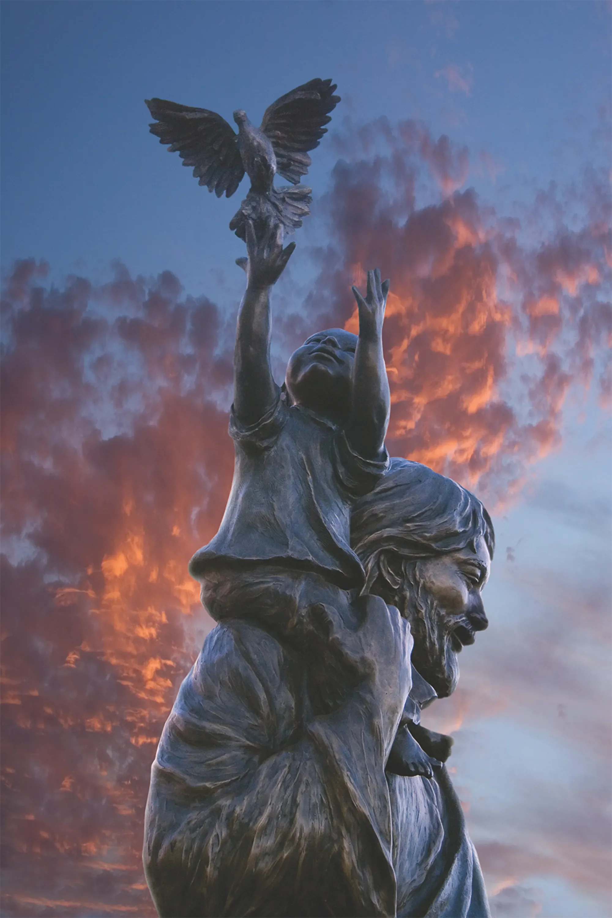 A bronze sculpture of a man with a beard, short hair, and a joyful expression, holding a child with outstretched arms reaching for a bird at sunset with a colorful sky in the background.