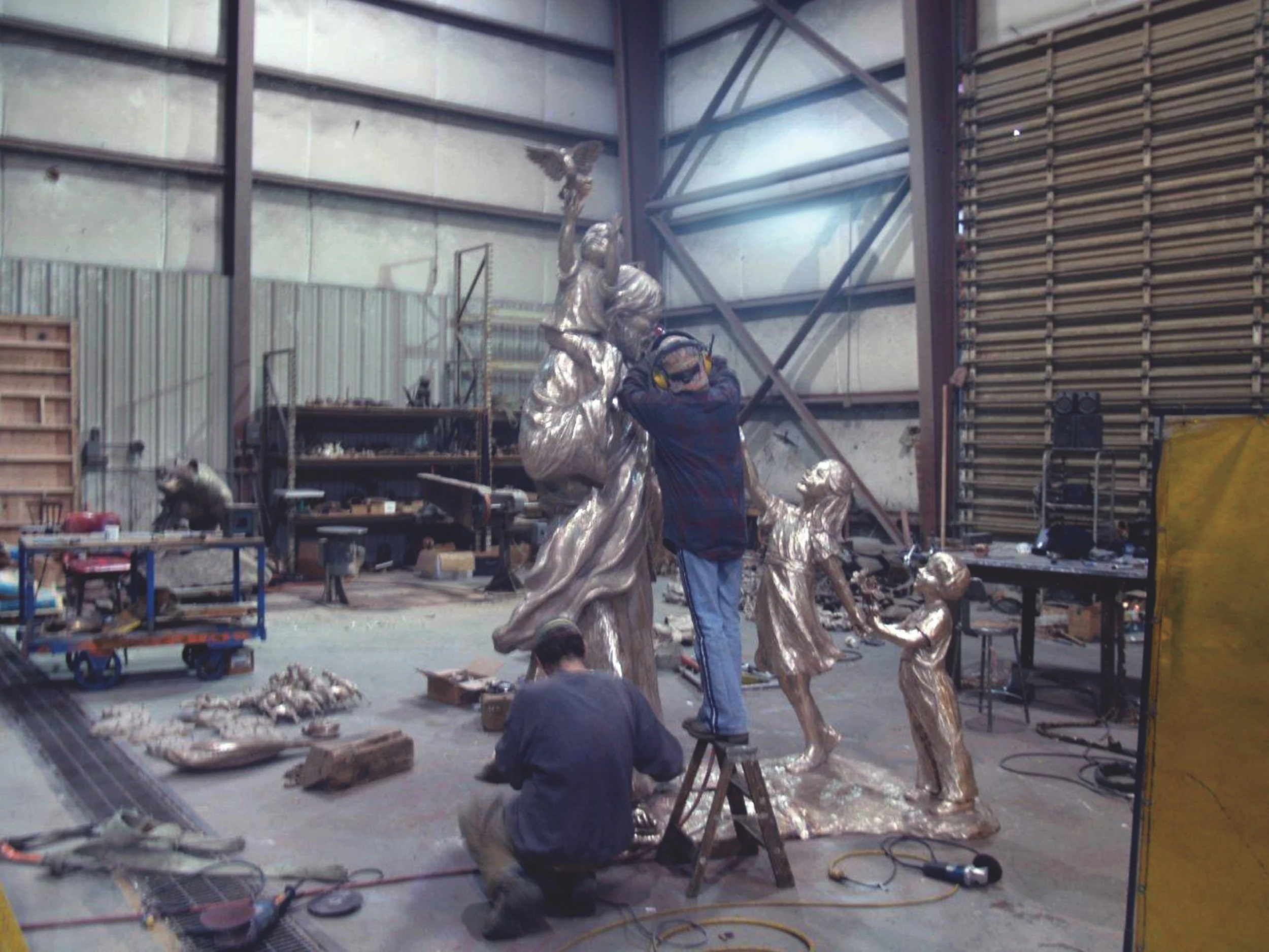 Workers in Rosalind's warehouse sculpture studio, working on metallic sculptures of children and angels, with tools and supplies scattered around.