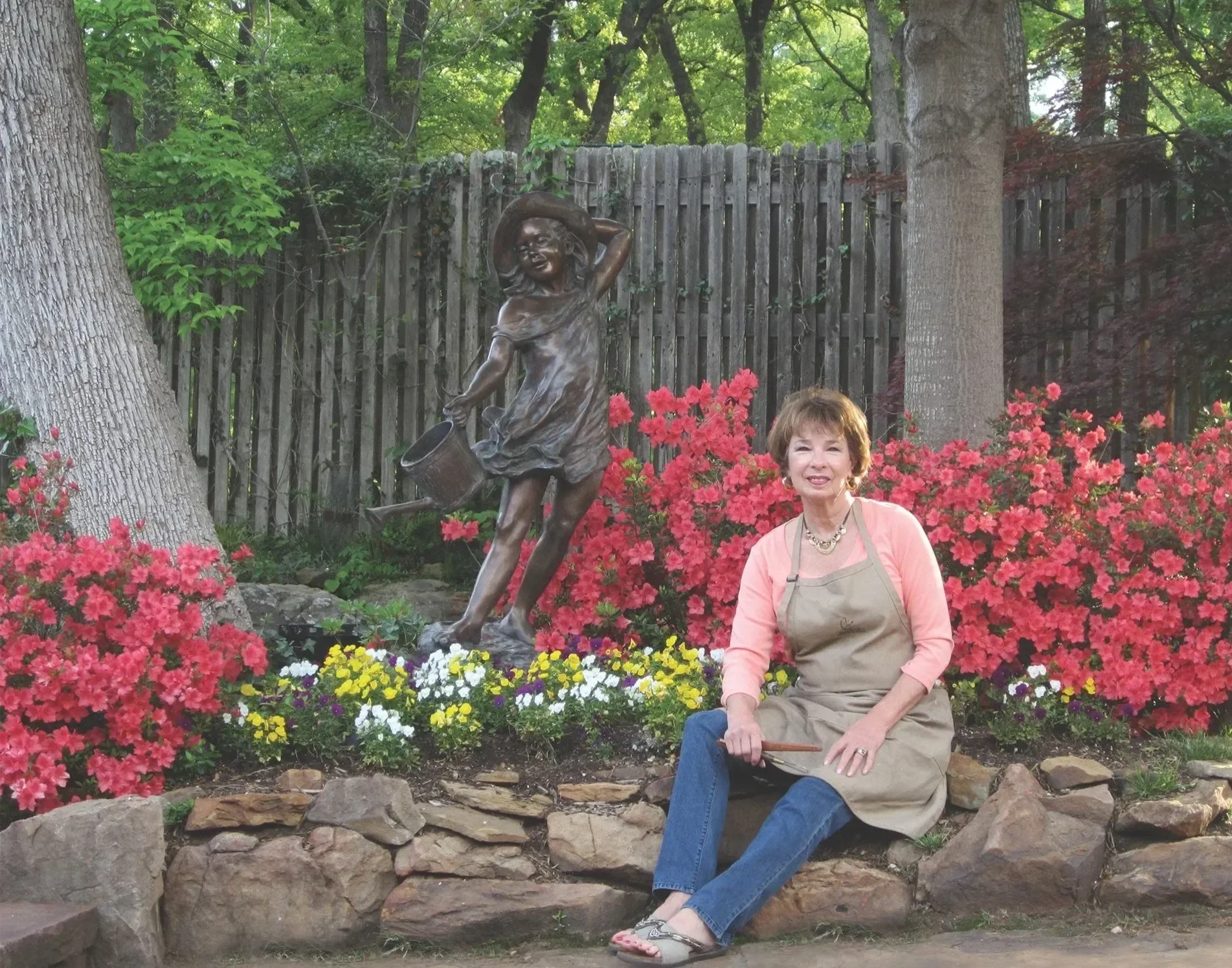 Rosalind Cook sitting on a rock, smiling, wearing a tan apron, pink shirt, blue jeans, and sandals, in a garden with pink azaleas, colorful flowers, trees, and a bronze statue of a girl watering plants in the background.