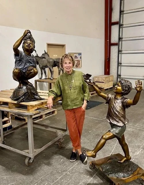 Rosalind Cook standing between two bronze sculptures of children, one with an outstretched arm and the other with a hand on her hip, in an industrial workshop with sculptures and supplies.