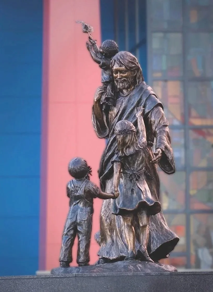 A bronze sculpture of Jesus Christ with three children, one holding his hand and another reaching out to him, with one child sitting on his shoulder and reaching tall.