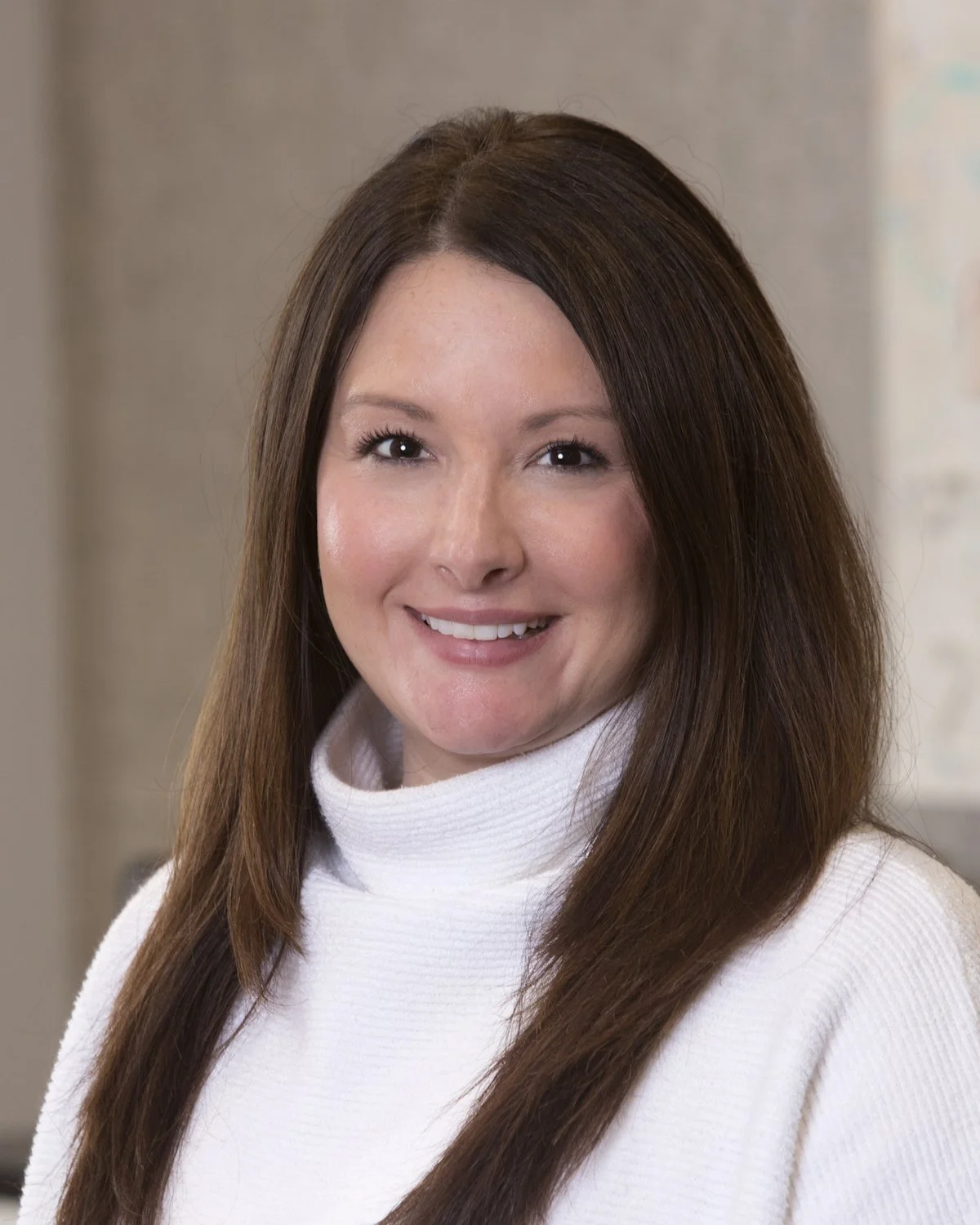 A woman with shoulder-length black hair, wearing a black top and brown blazer, smiling in front of a wall with framed artwork.