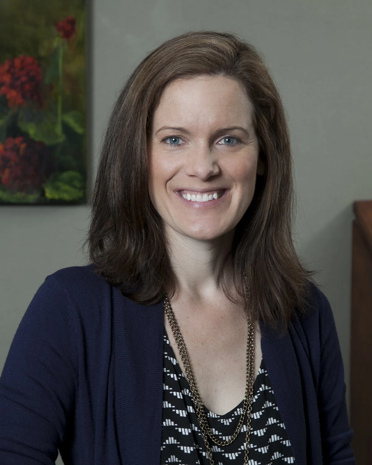 A woman with shoulder-length brown hair, smiling, wearing a navy blue cardigan over a patterned top, standing in front of a blurred background with colorful artwork.