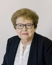 A portrait of an older woman with short curly hair, glasses, wearing a dark blazer over a white blouse, against a plain background.