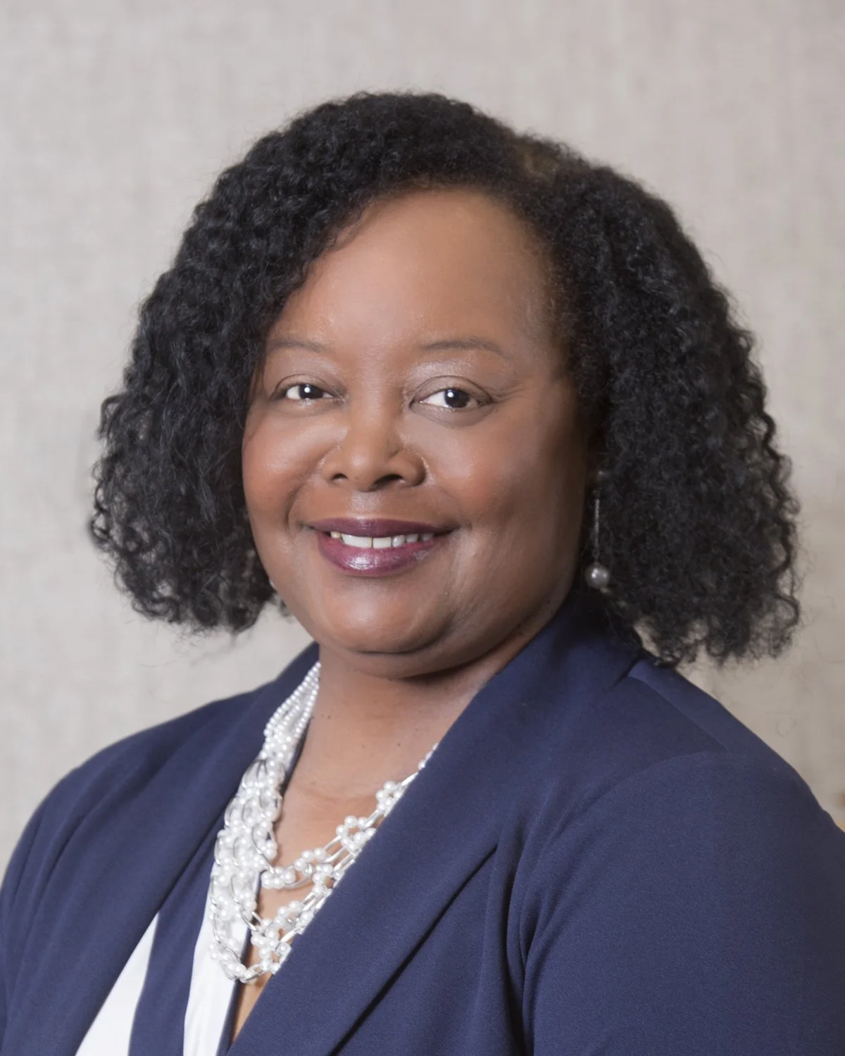 A smiling woman with gray hair wearing a light-colored blazer and pearl necklace, standing indoors.