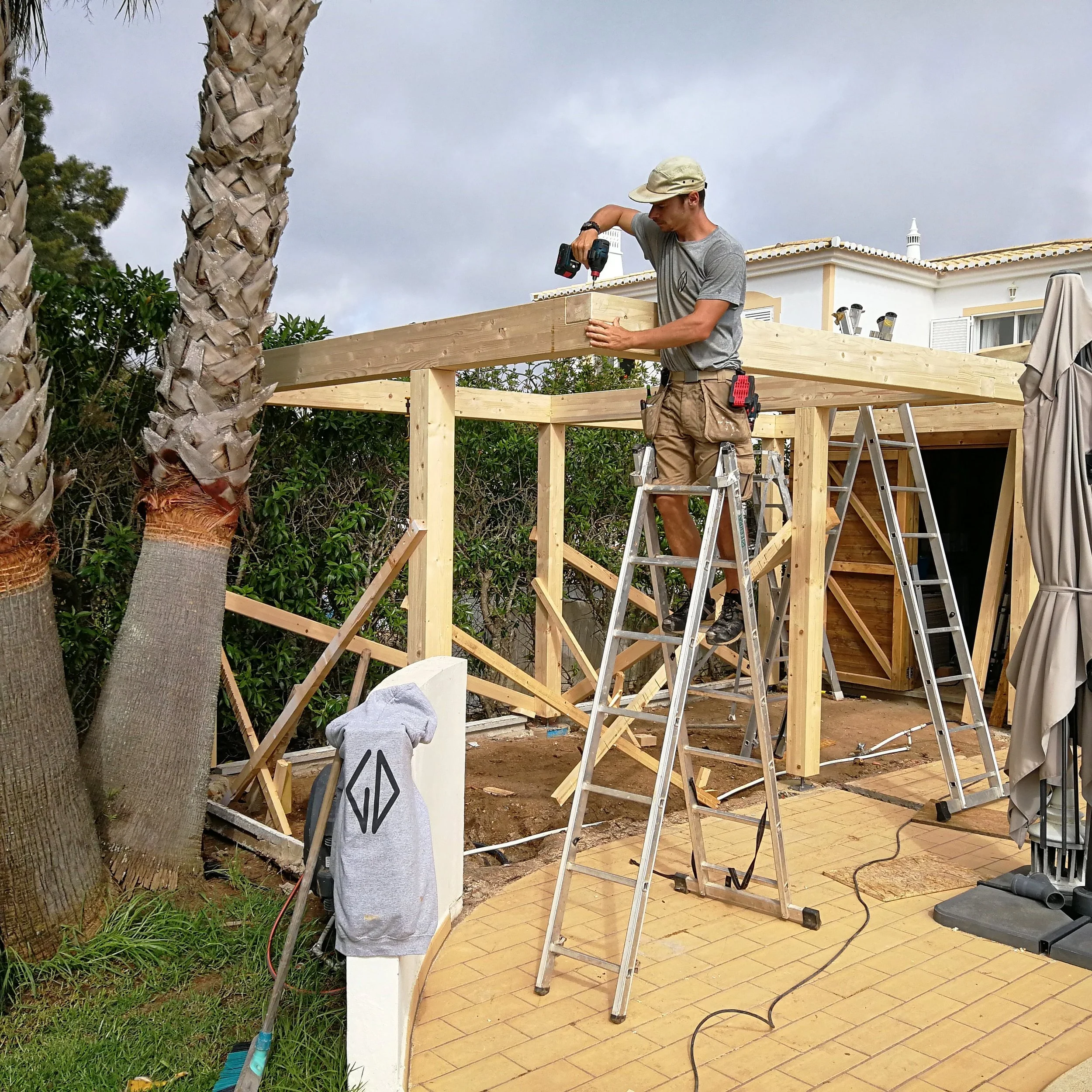A man wearing a tan cap and gray t-shirt is working on a construction project outdoors, using a drill on a wooden beam. He stands on a ladder next to a partially built wooden deck, with a house in the background and a cloudy sky above.