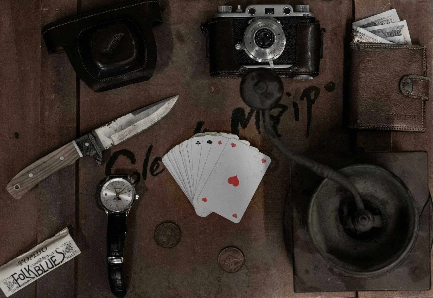 A collection of items on a wooden surface including a vintage camera, a deck of playing cards, a pocket wallet with cash, a wristwatch, a technical knife, a black leather case, and a glass with a beverage. There are also some coins and a handwritten note that says 'love'.