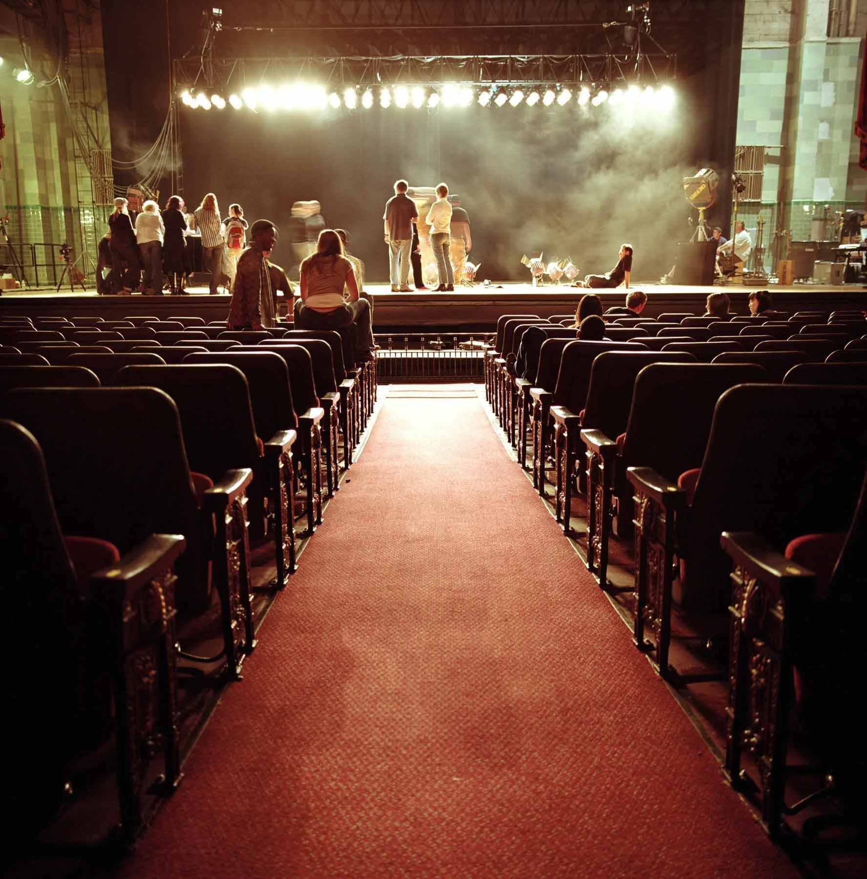 Stage set up in an auditorium with rows of chairs, stage lighting, and a few people waiting or preparing for a performance.